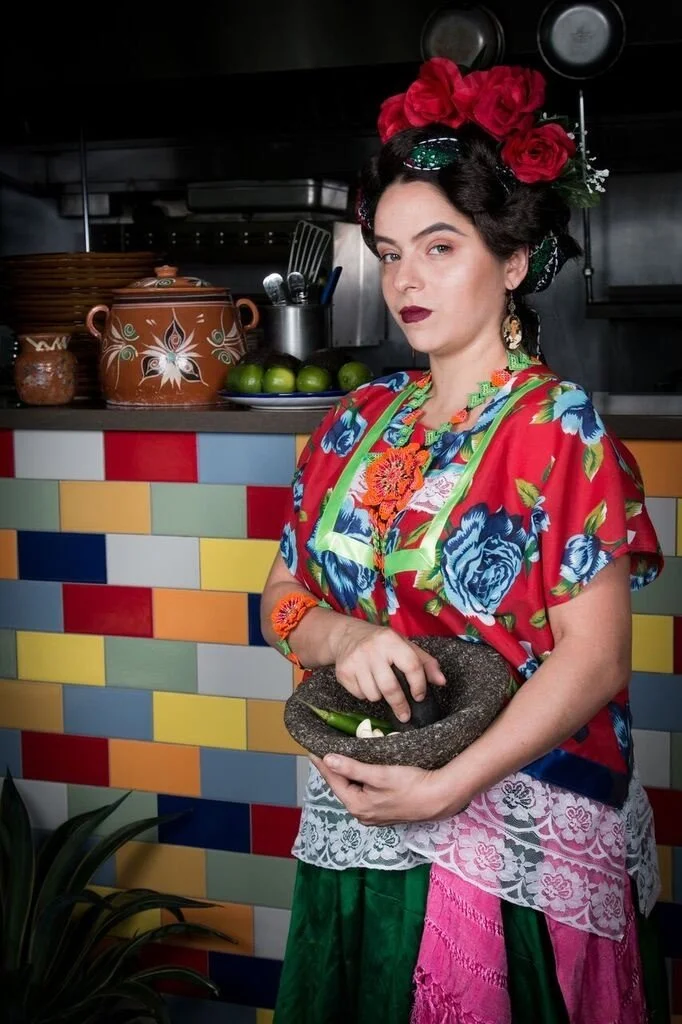 Woman in traditional Mexican attire holding a mortar and pestle in a colorful kitchen with tiled wall and pottery.