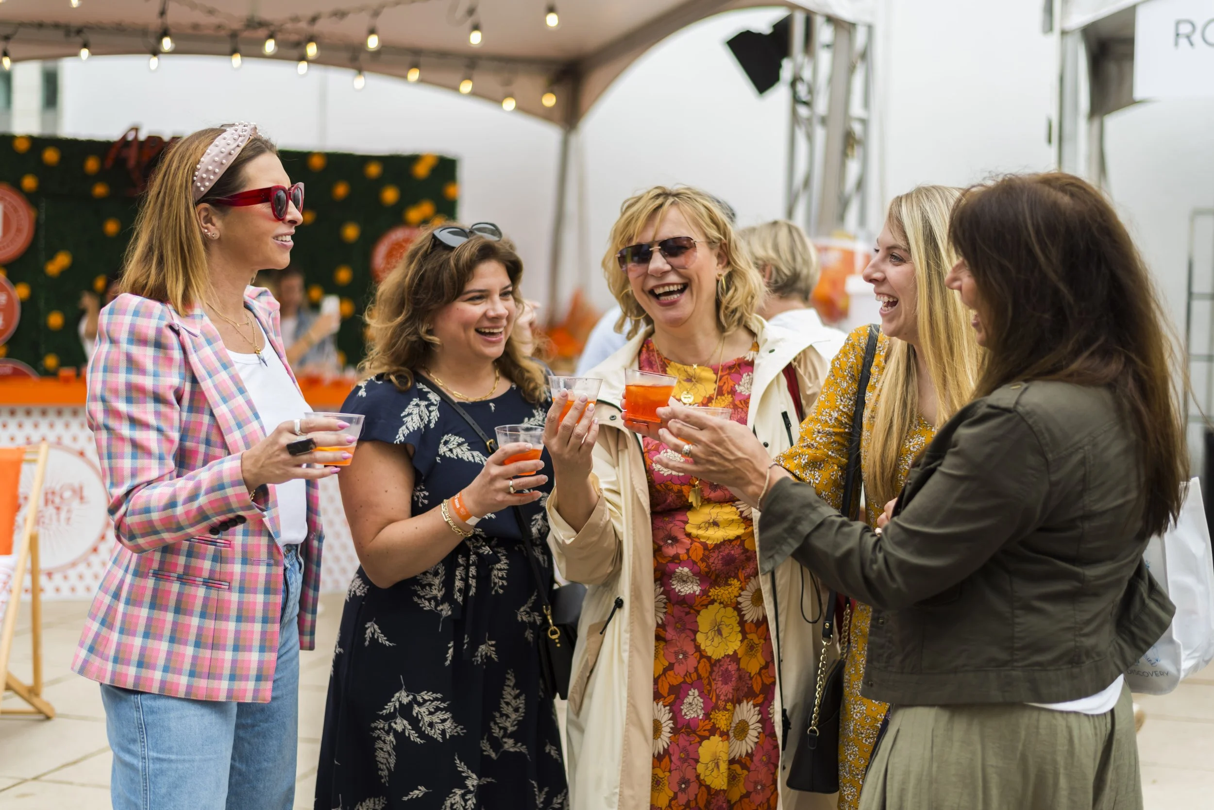 A group of women smiling and chatting at Chicago Gourmet, holding drinks in disposable cups, under a tent with string lights.