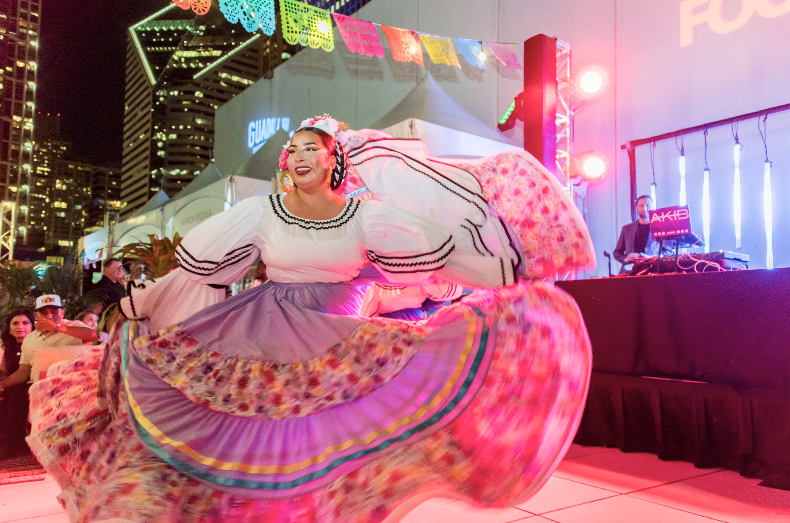 A woman in traditional Mexican clothing twirls at a cultural event or celebration, with a colorful skirt and embroidered blouse, near a DJ booth with city buildings in the background.