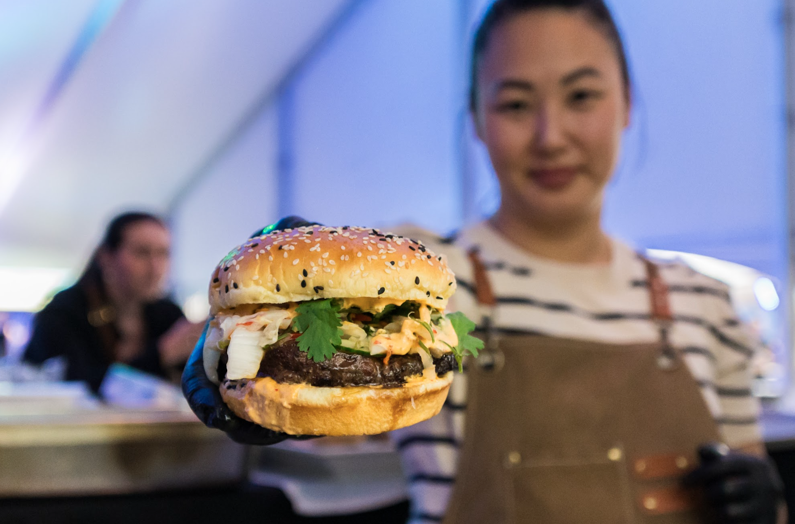 A woman holding a burger with sesame seed bun, lettuce, cilantro, and burger patty at an indoor event.