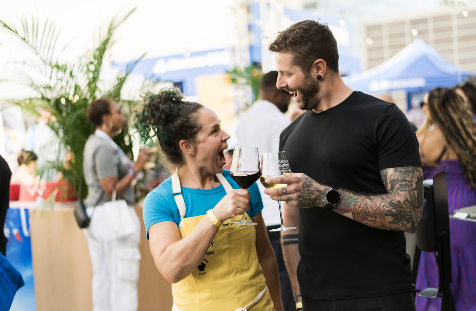 Two people, a woman with curly hair in a blue shirt and a yellow apron and a man with tattoos and a black shirt, are smiling and holding glasses of red wine while talking outside