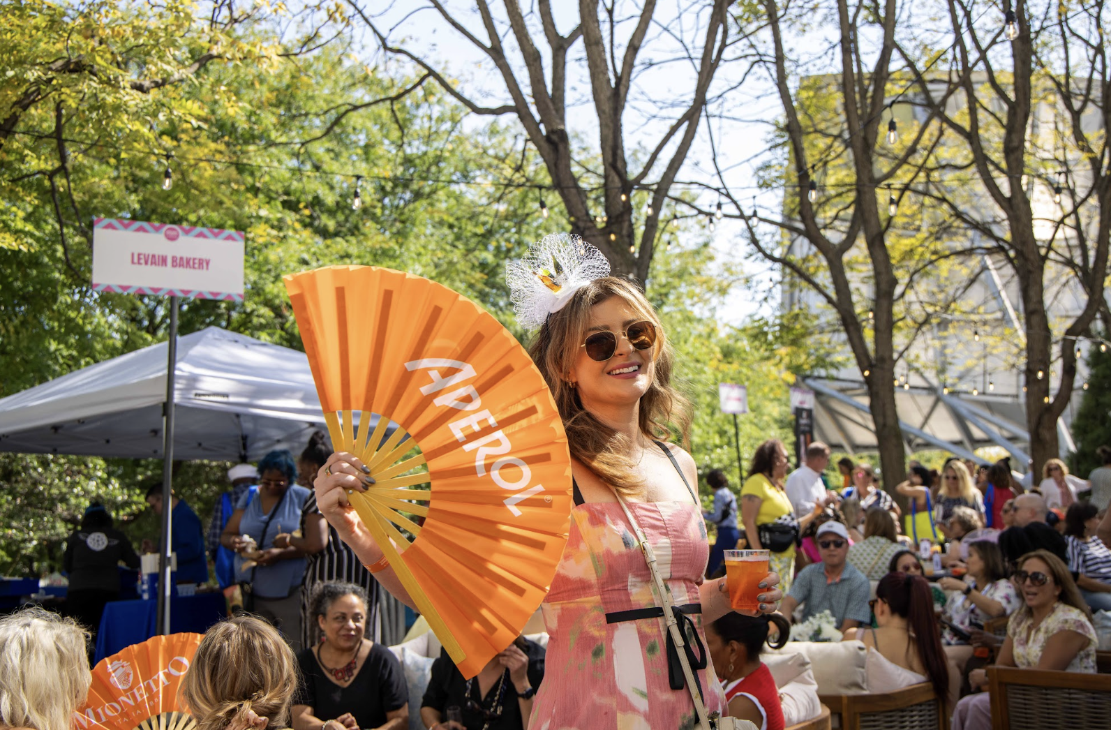Woman wearing sunglasses and a floral dress smiling at outdoor event, holding orange fan with