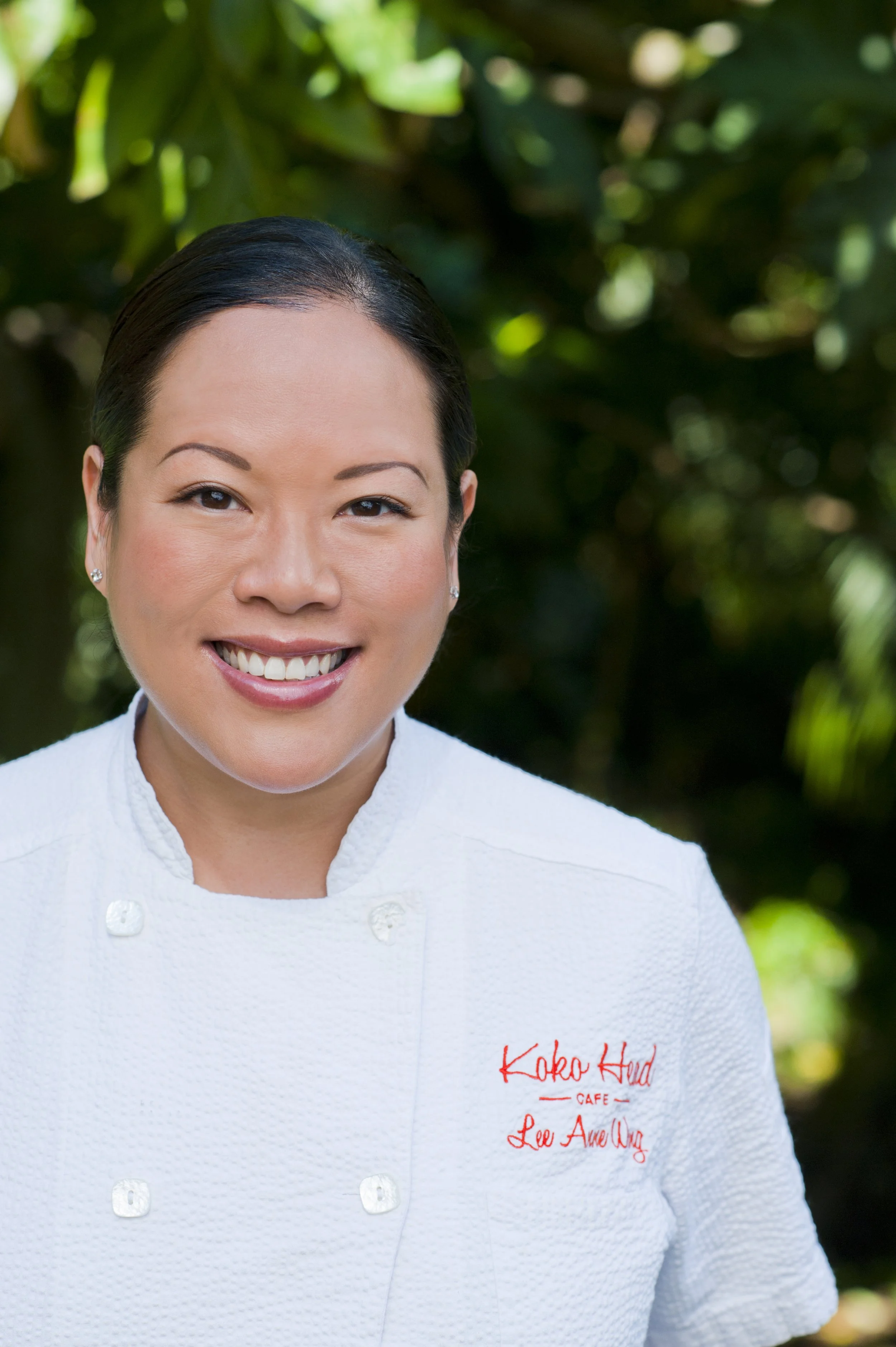 Chef Lee Ann Wong wearing a white textured chef coat with red embroidery on the left side that reads 'Koko Head Cafe' and 'Lee Anne Wong'. The background is blurred greenery.