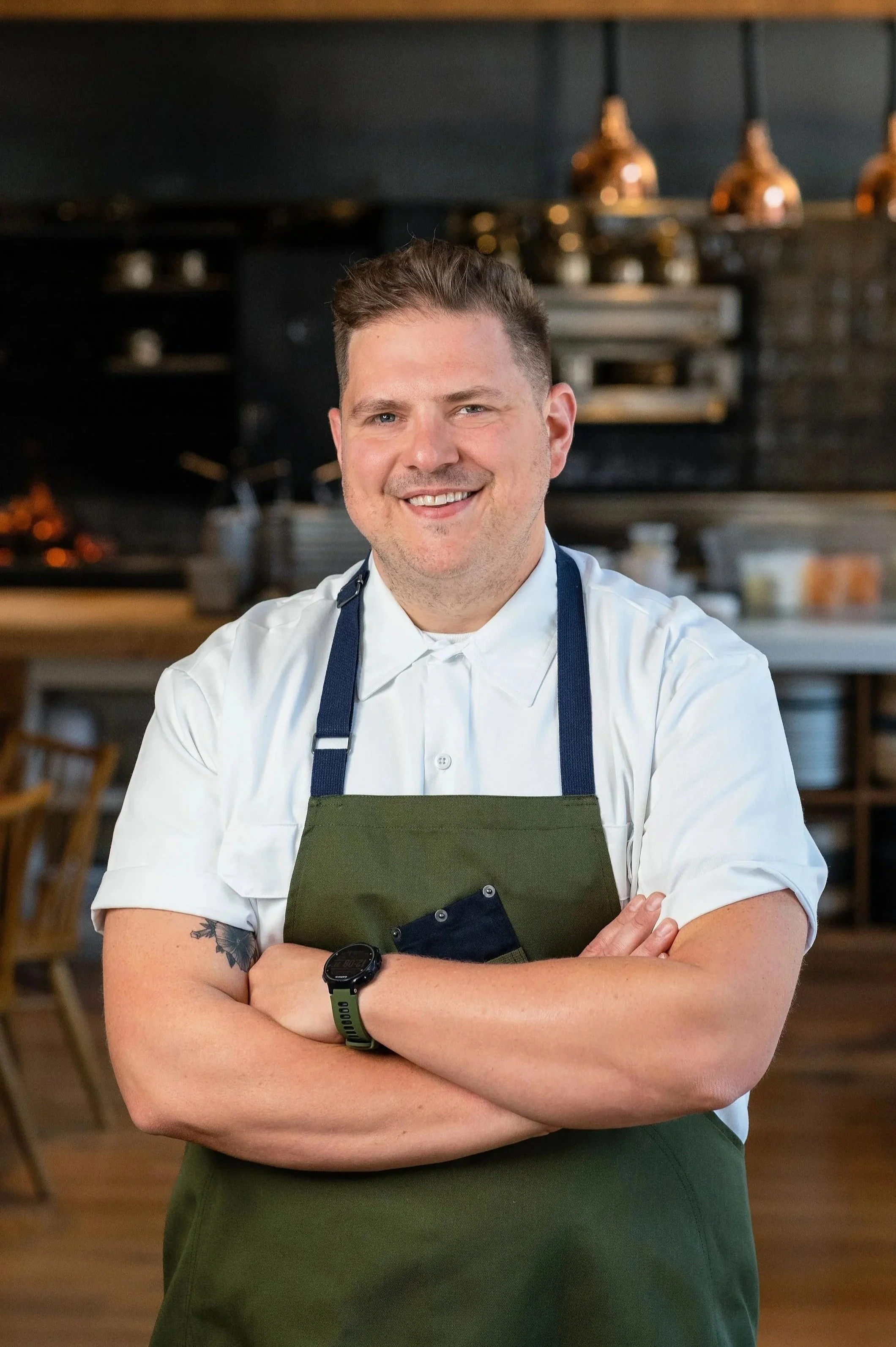 Chef Joe Flamm is standing with crossed arms, wearing a white chef's coat and a green apron, in a restaurant kitchen with a dark background.