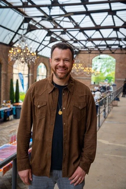 A smiling man with a beard and short hair standing indoors in a brick building with a glass ceiling, string lights, and tables in the background.