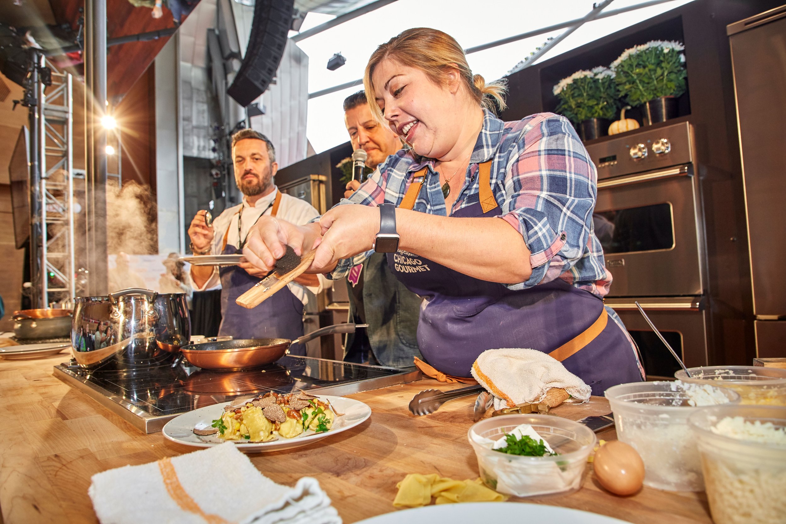A woman cooking in a restaurant kitchen, surrounded by ingredients, with two men watching her. She is smiling and holding a cooking utensil, wearing a plaid shirt and apron. The background shows kitchen appliances and decorative plants.
