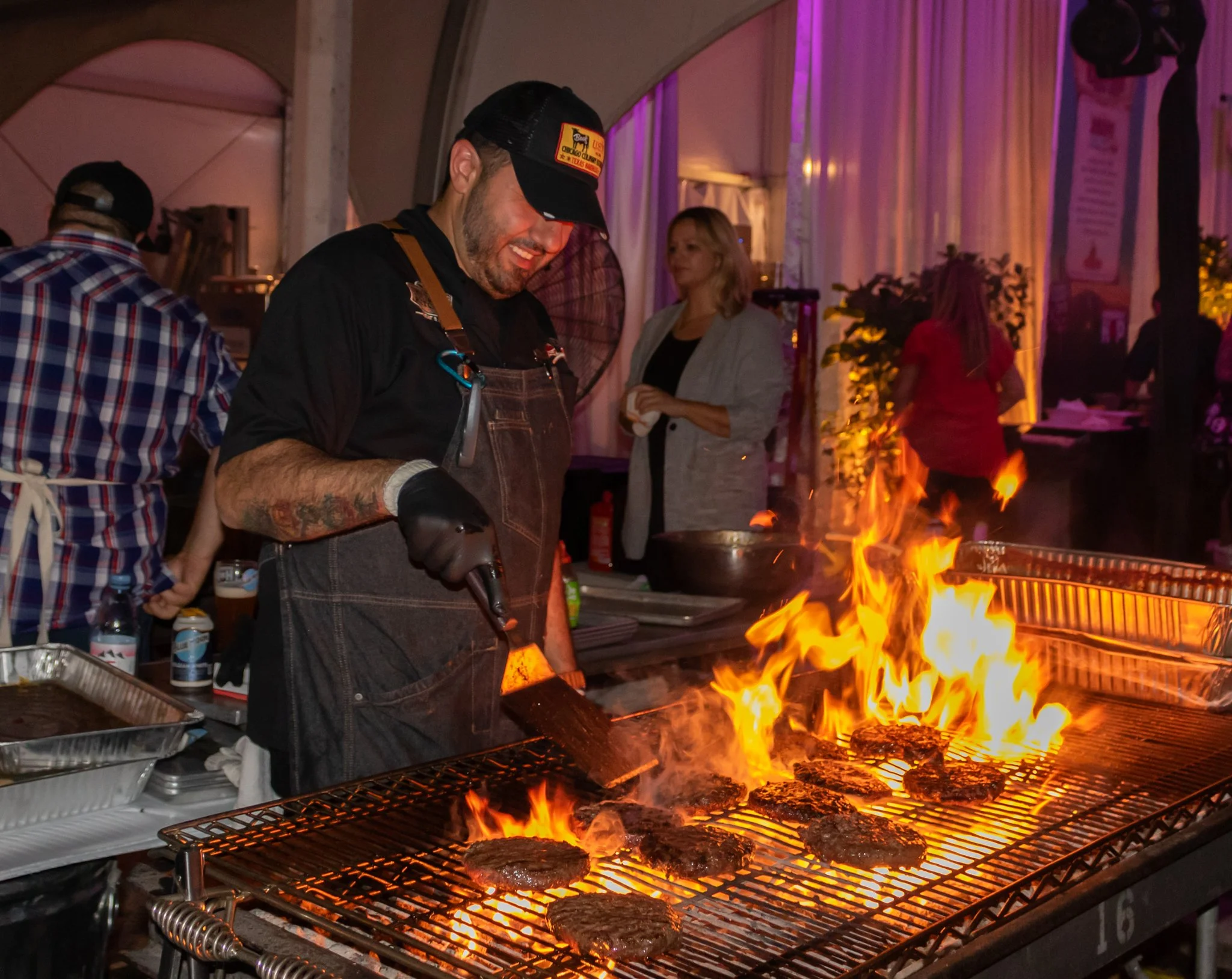 A man cooking steaks on an open flame grill with other people standing around in a tented or covered area with purple and blue lighting.