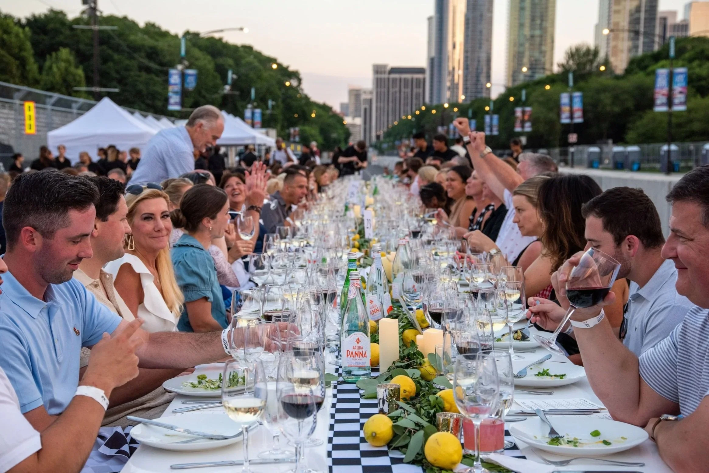 Long outdoor dinner party with people seated along a table, decorated with lemons, candles, and greenery, with city buildings in the background during evening.
