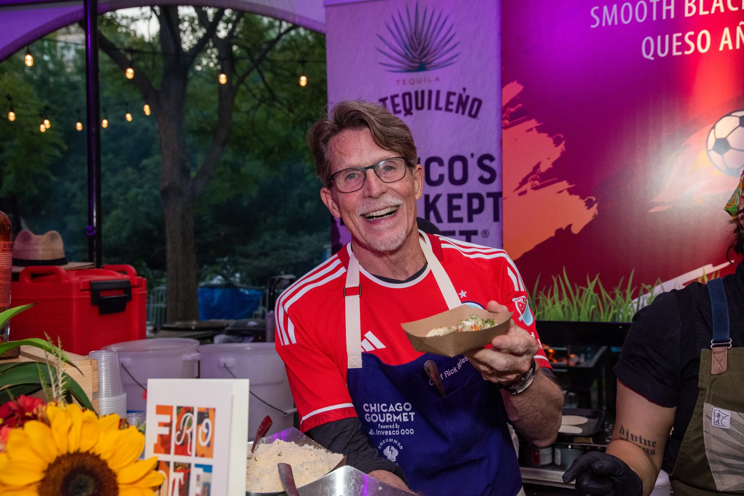 Chef Rick Bayless wearing a red and white striped shirt holding a bowl of food, smiling at an outdoor event with trees and colorful banners in the background.