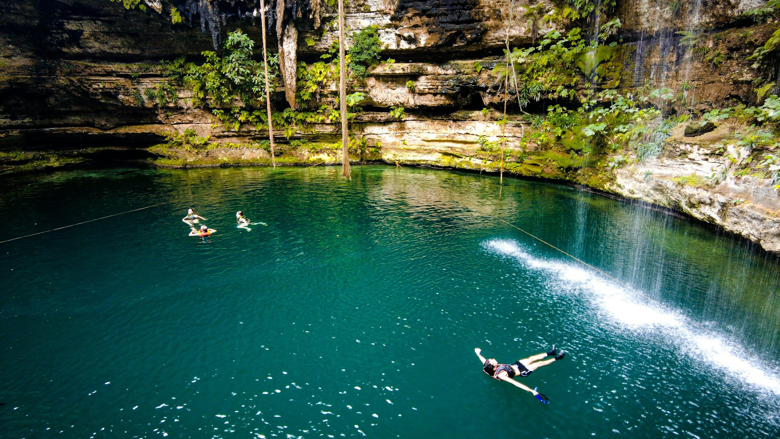 People swimming and snorkeling in a natural water-filled cave with lush greenery and waterfall features.