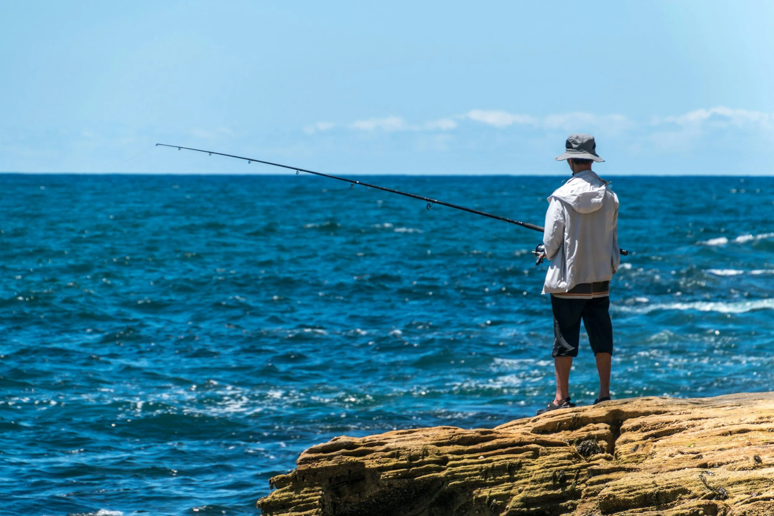 A person fishing on a rocky shoreline, wearing a wide-brimmed hat, light jacket, and shorts, overlooking the blue ocean with a partly cloudy sky.