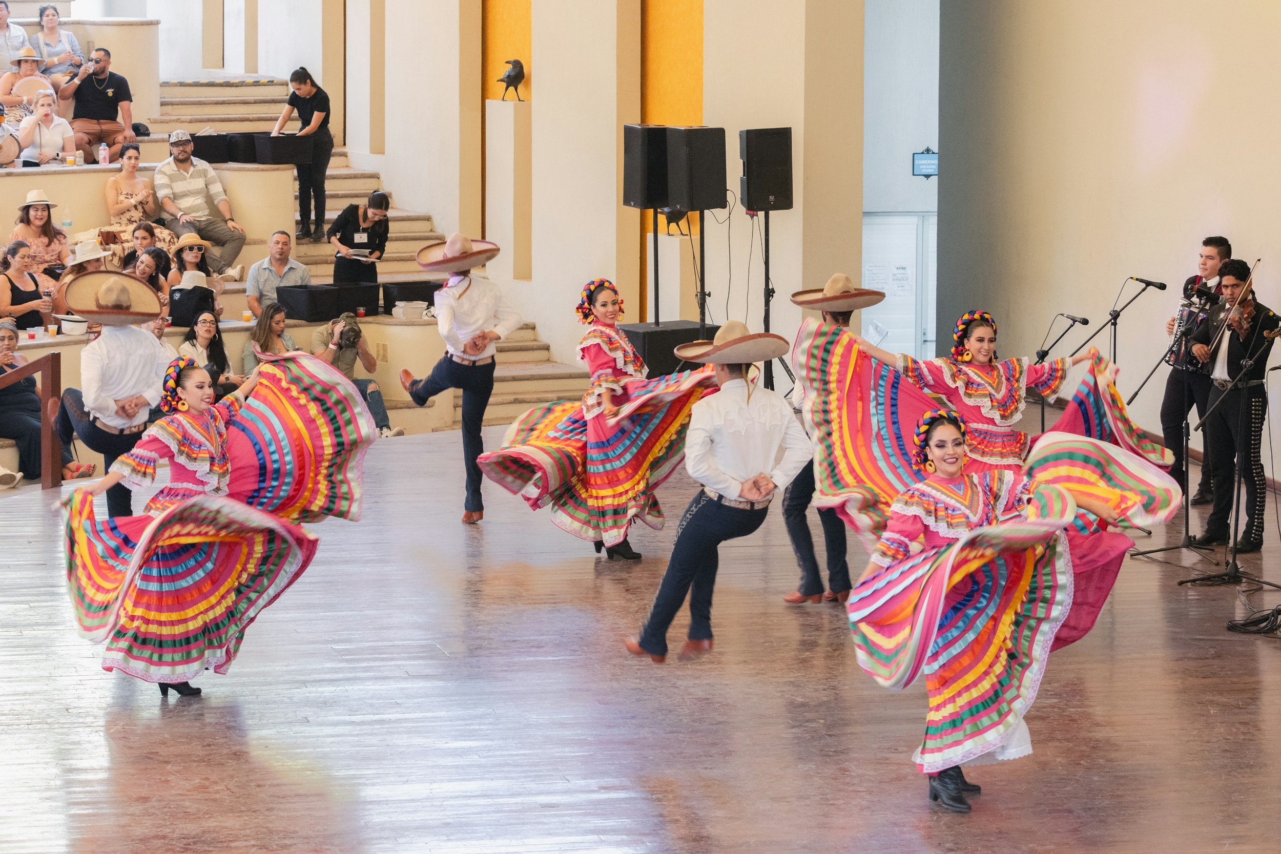 Group of dancers performing traditional Mexican folkloric dance in colorful dresses and sombreros, with an audience watching nearby.