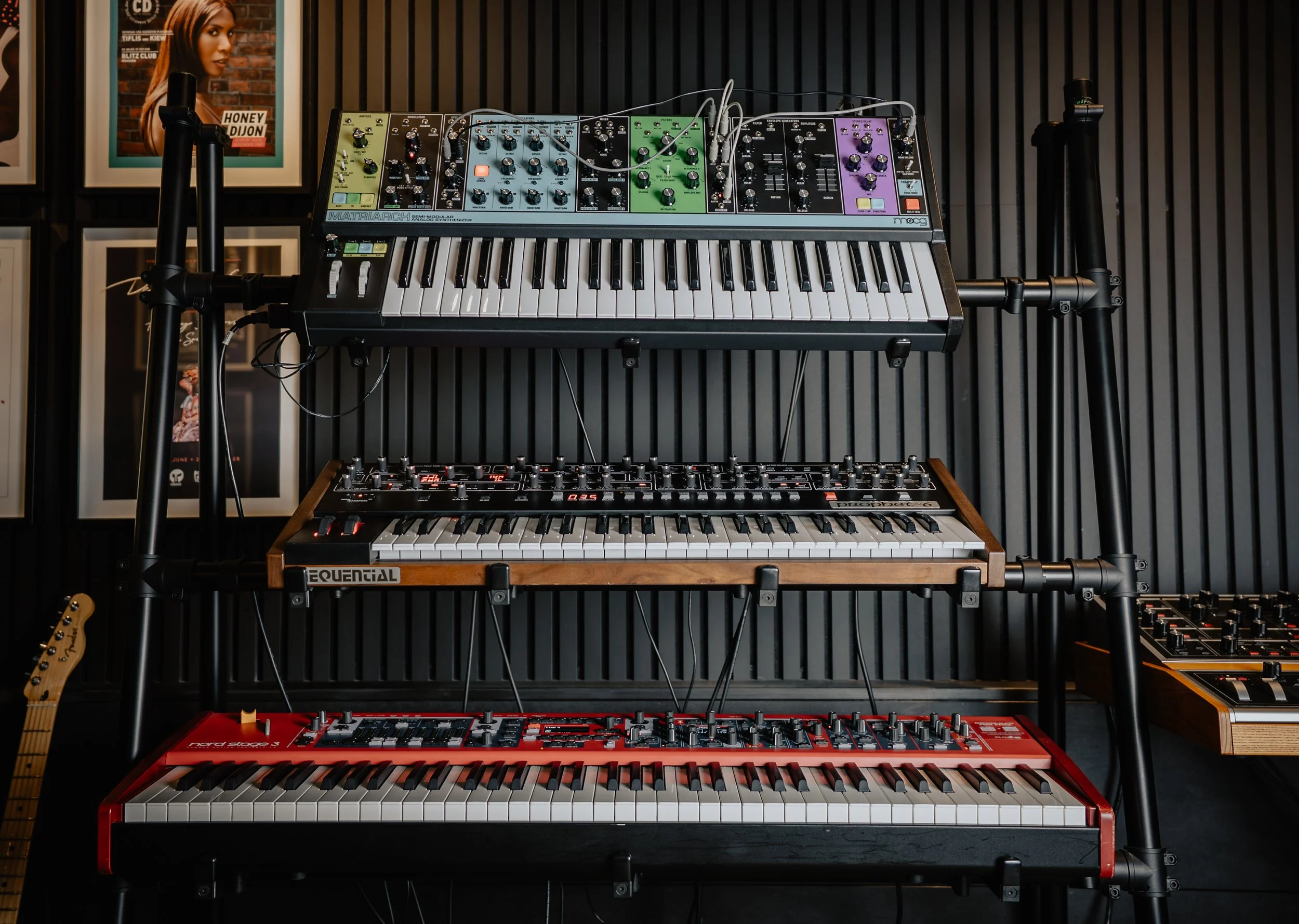 A music studio setup with three levels of synthesizers on a black metal and wood rack. The top level features a Moog Matriarch synthesizer, the middle level has a Sequential Prophet synthesizer, and the bottom level contains a Nord Stage 3 keyboard. To the right, there is a small section of another synthesizer with various knobs.