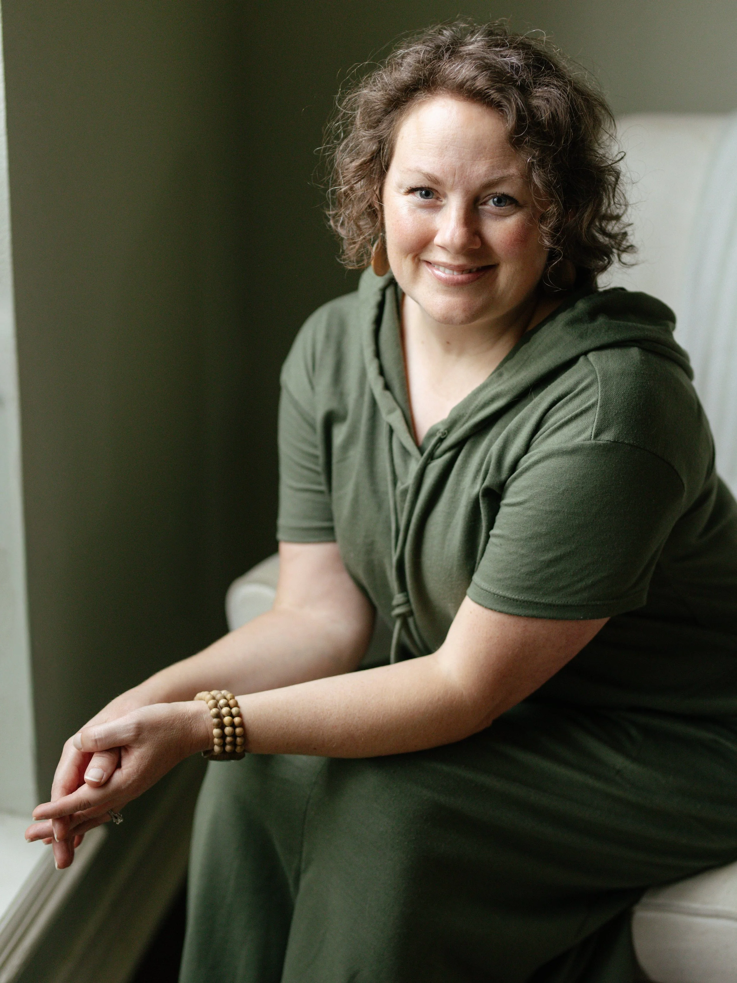 A woman with short, curly brown hair, wearing beaded bracelets and earrings, dressed in an olive green hoodie, sitting by a window and smiling.