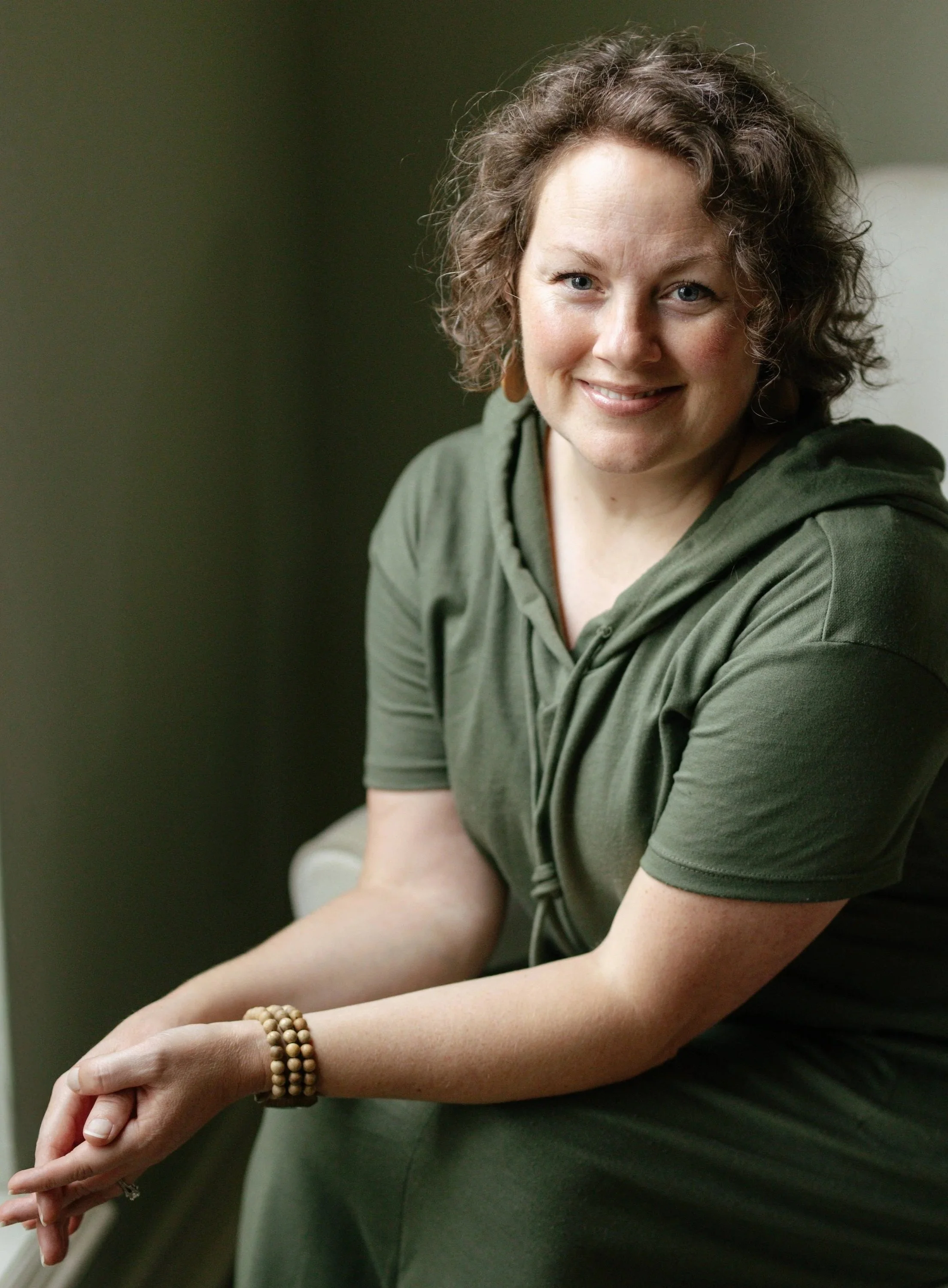 A woman with curly brown hair and blue eyes, wearing a green hoodie, sitting indoors with her hands resting on her arm, wearing beaded bracelets, smiling softly at the camera.