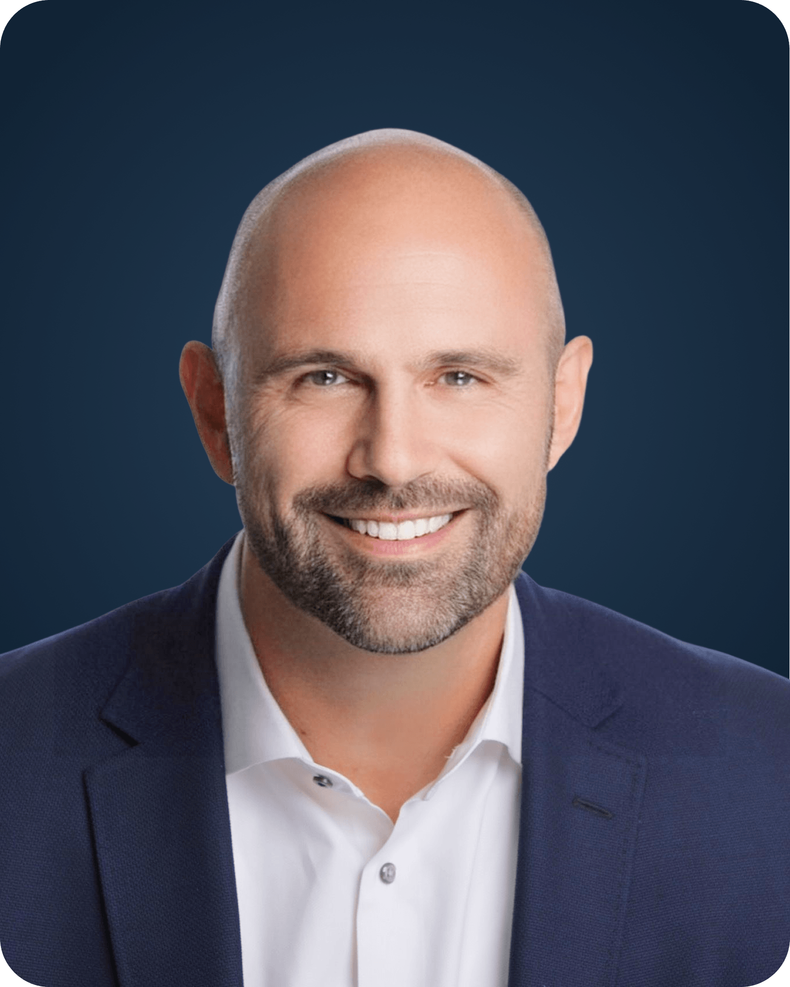Close-up of a smiling man with a shaved head wearing a navy blue suit and white shirt against a dark background.