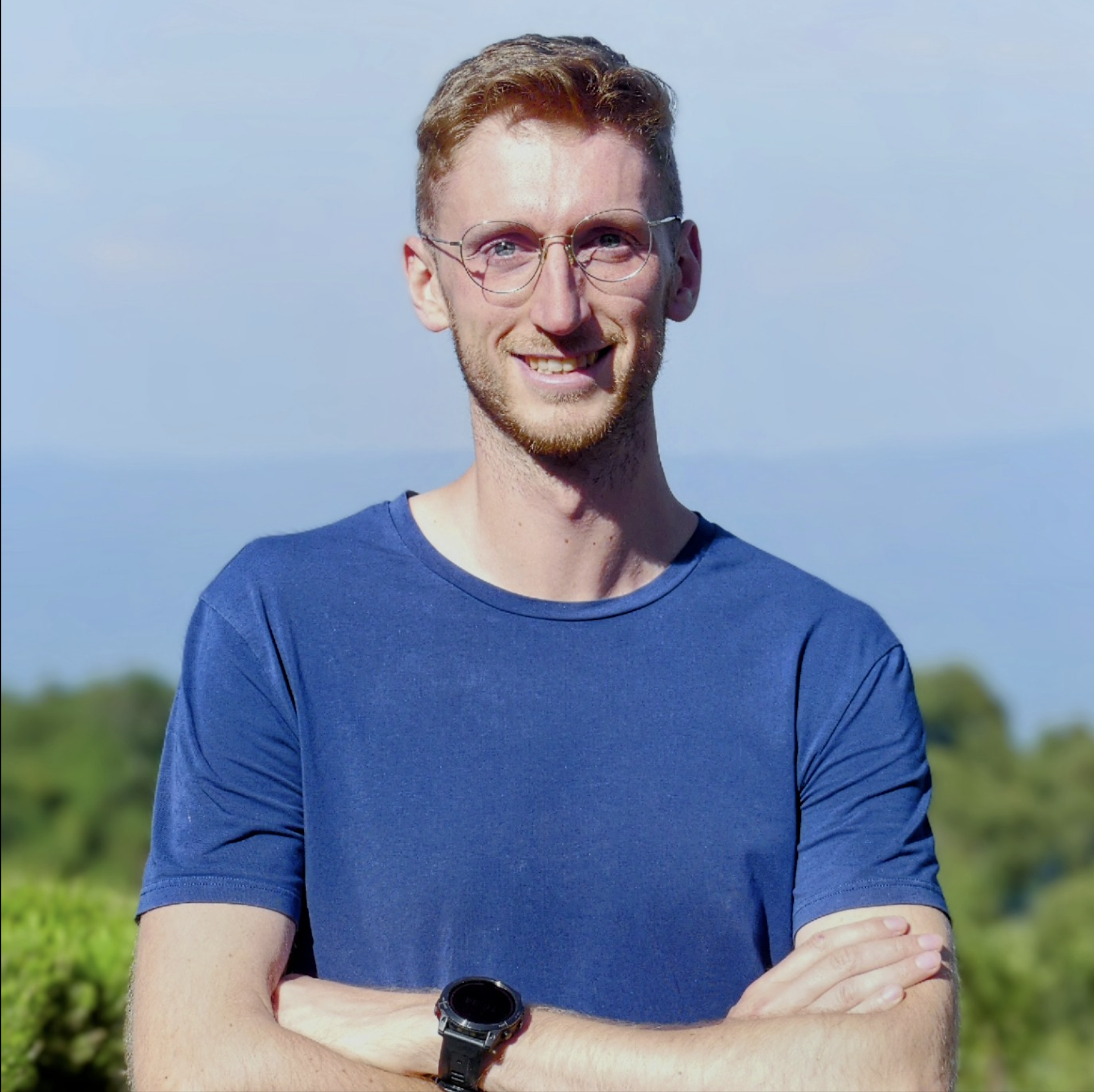 A young man with glasses and a beard, smiling, standing outdoors with arms crossed, wearing a blue t-shirt and a watch, with a blurred green landscape and blue sky in the background.