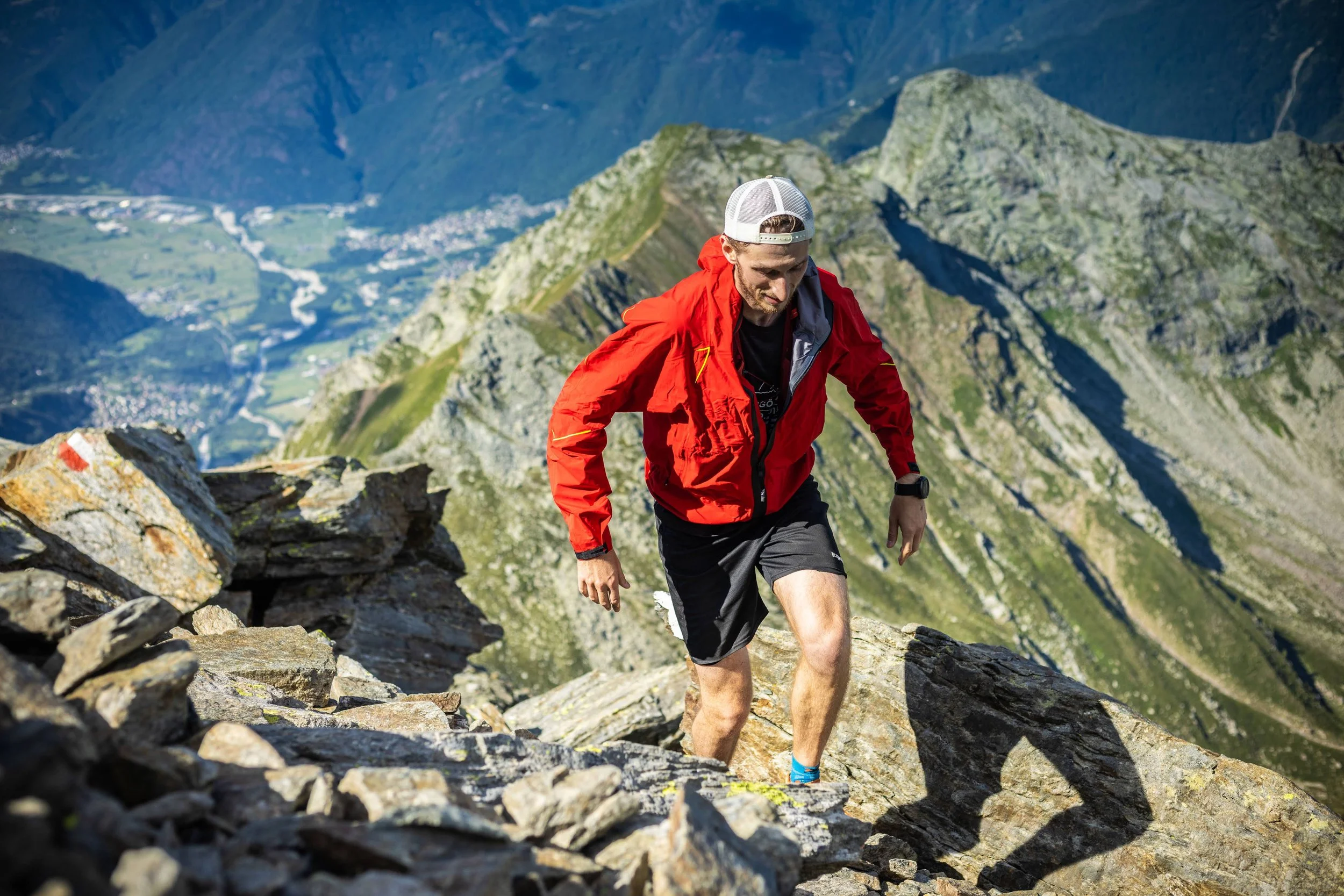 A man in athletic gear, including a red jacket, black shorts, and a white cap, hikes up a rocky mountain trail with mountain peaks and valleys in the background.
