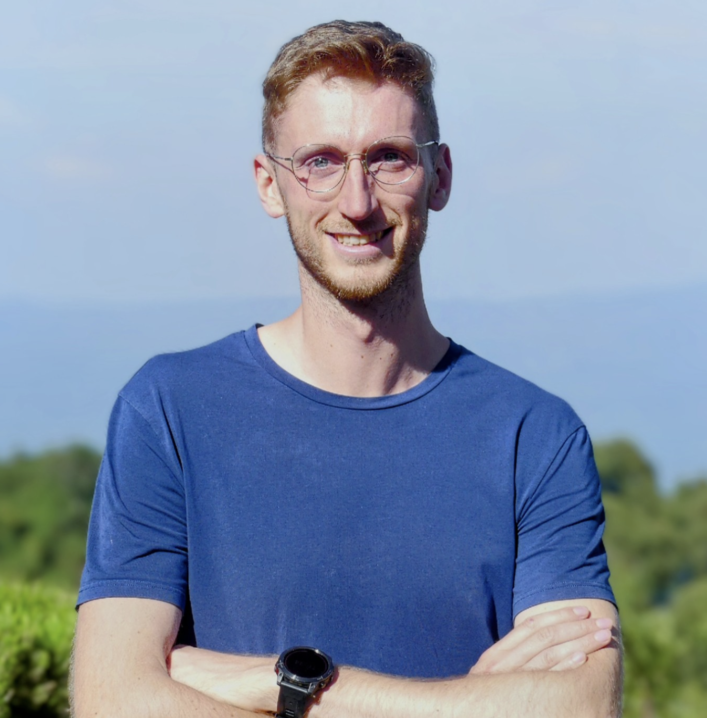 A young man with glasses and a beard, smiling, standing outdoors with arms crossed, wearing a blue t-shirt and a watch, with a blurred green landscape and blue sky in the background.