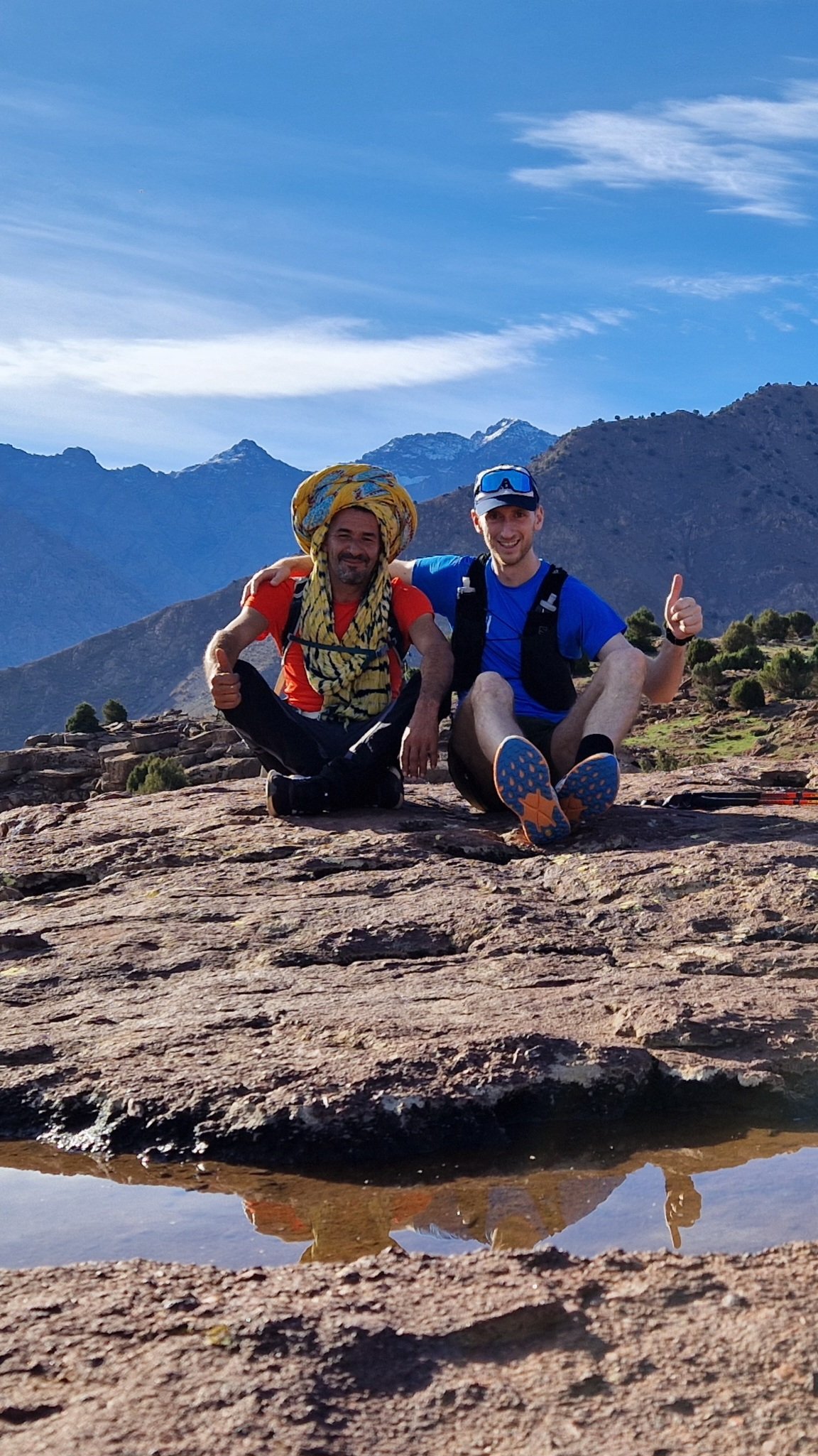 Two smiling men sitting on a rocky surface outdoors with Mountains and blue sky in the background, one wearing a yellow headscarf, red shirt, and the other in a blue shirt with a backpack.