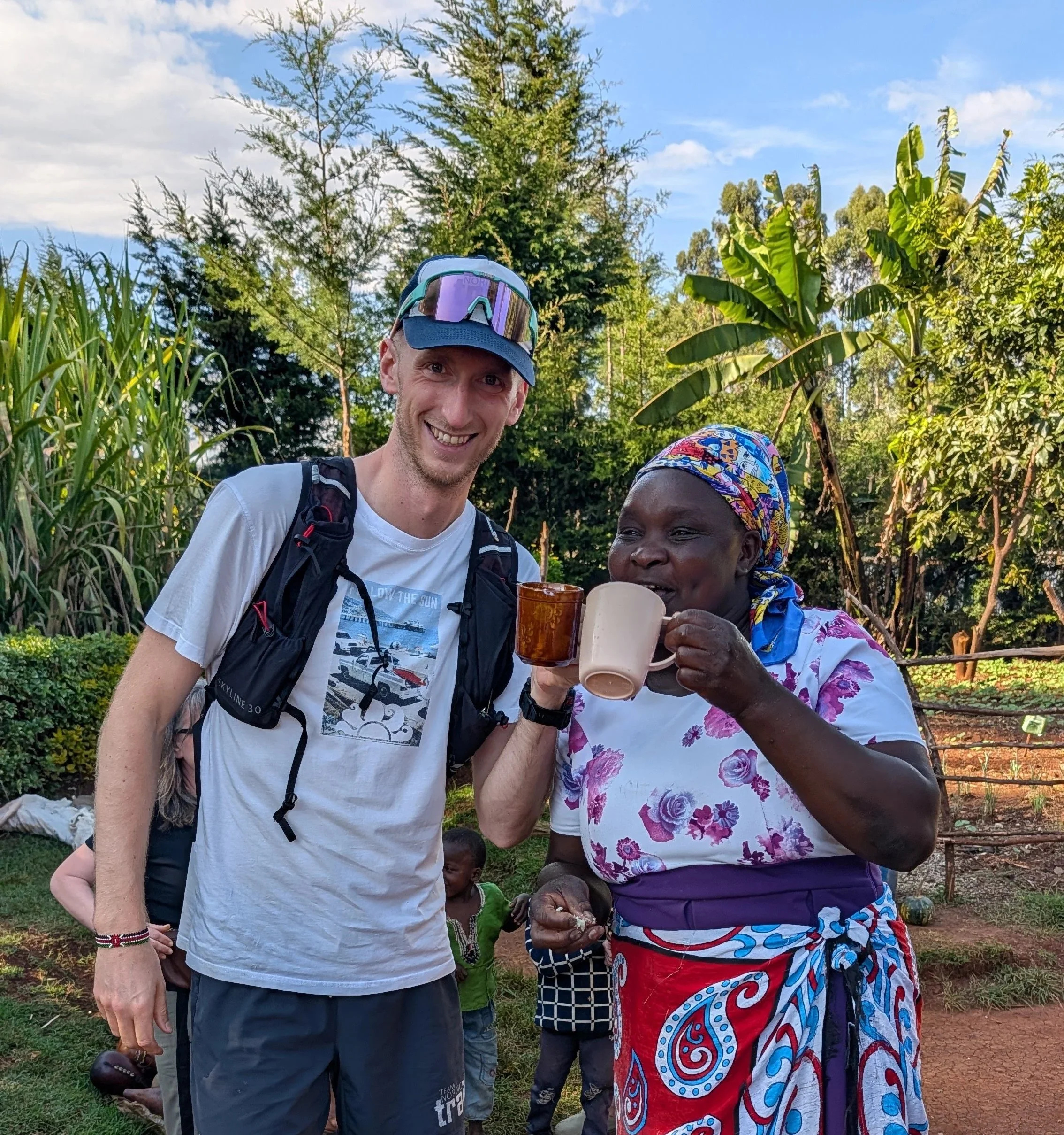 A man and a woman smiling and holding coffee cups outdoors in a lush garden with trees in the background, some children visible behind them.