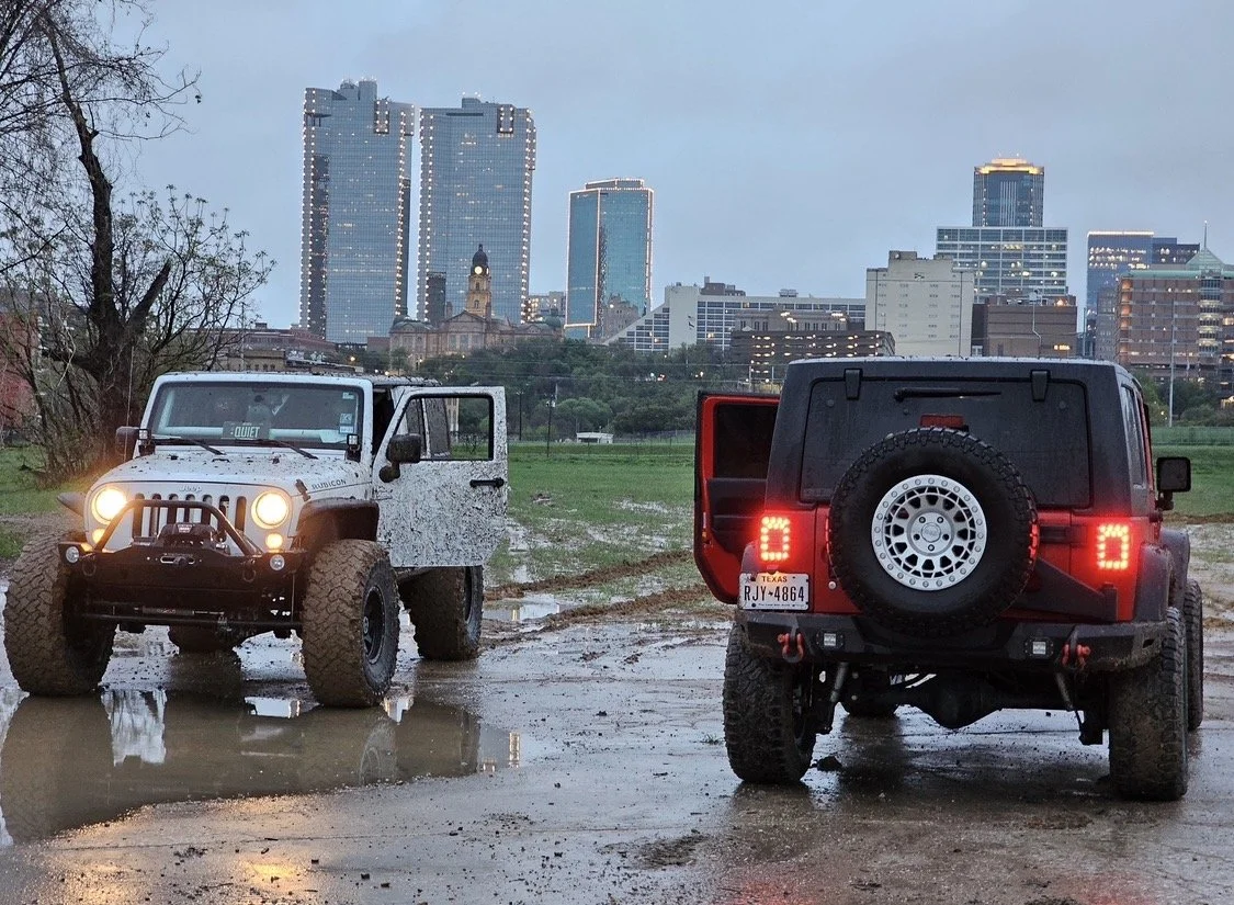 Two off-road vehicles parked on a muddy field with puddles, with a city skyline in the background.