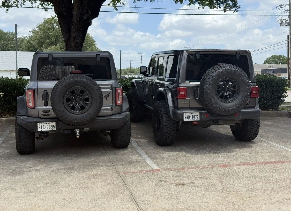 Two black and gray Jeep vehicles parked side by side in a parking lot, with spare tires mounted on their backs.