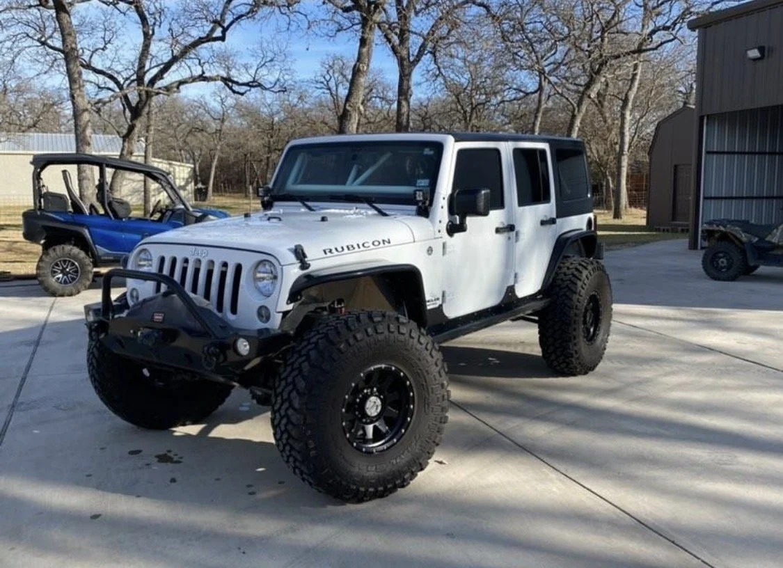 A white Jeep Wrangler Rubicon with large off-road tires and a black front bumper parked on a driveway, with trees and a building in the background.