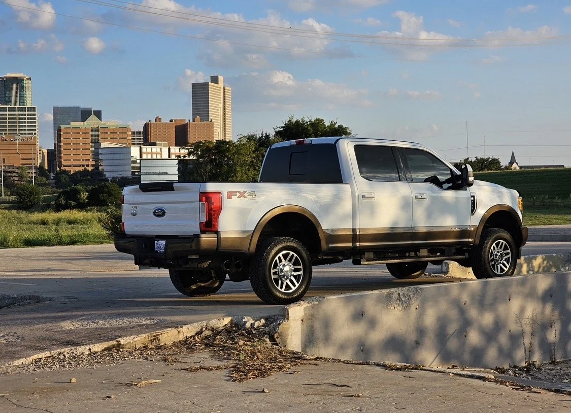 White Ford pickup truck with brown accents parked on a concrete surface against a city skyline with tall buildings, green grass, and a partly cloudy sky.