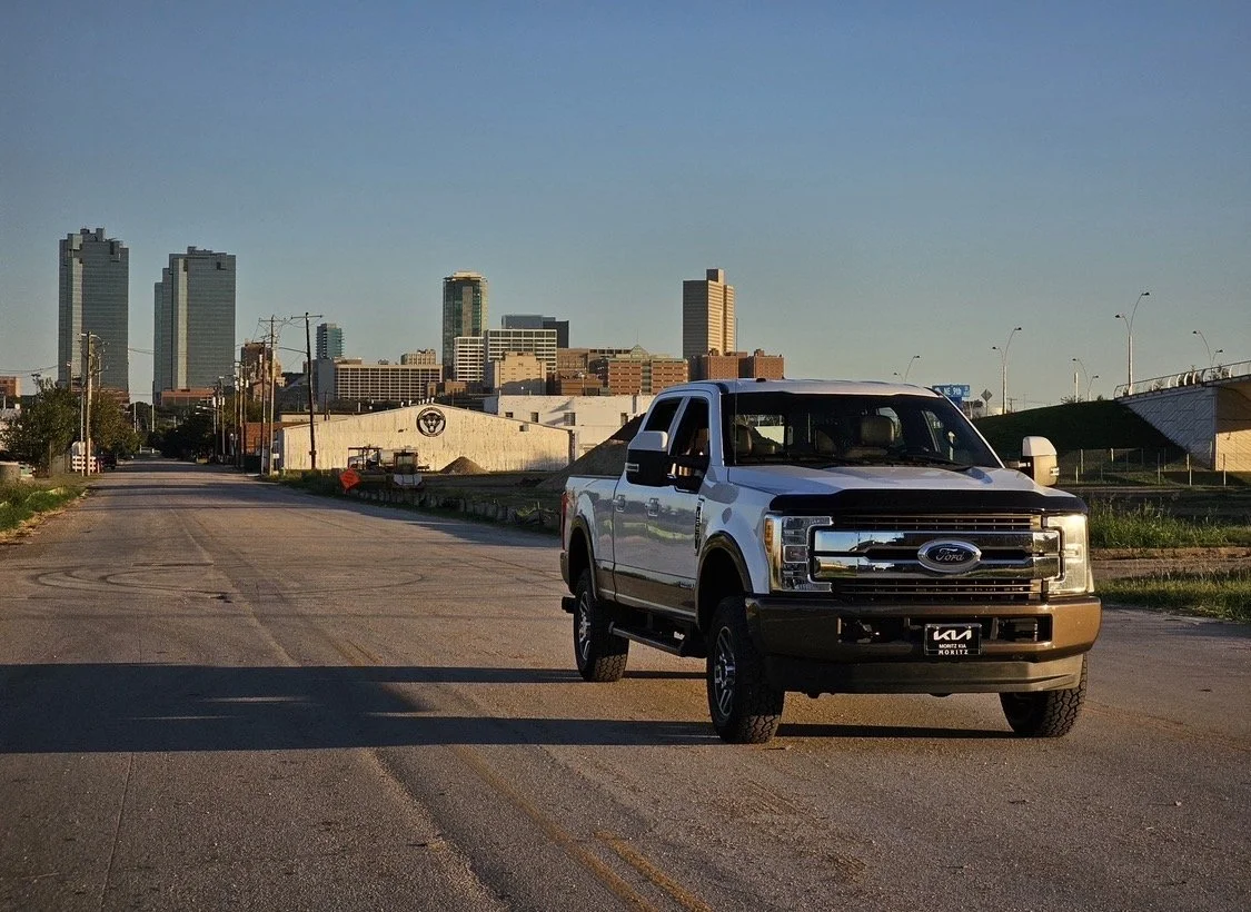 A white Ford pickup truck parked on an empty city street with skyscrapers in the background during sunset.
