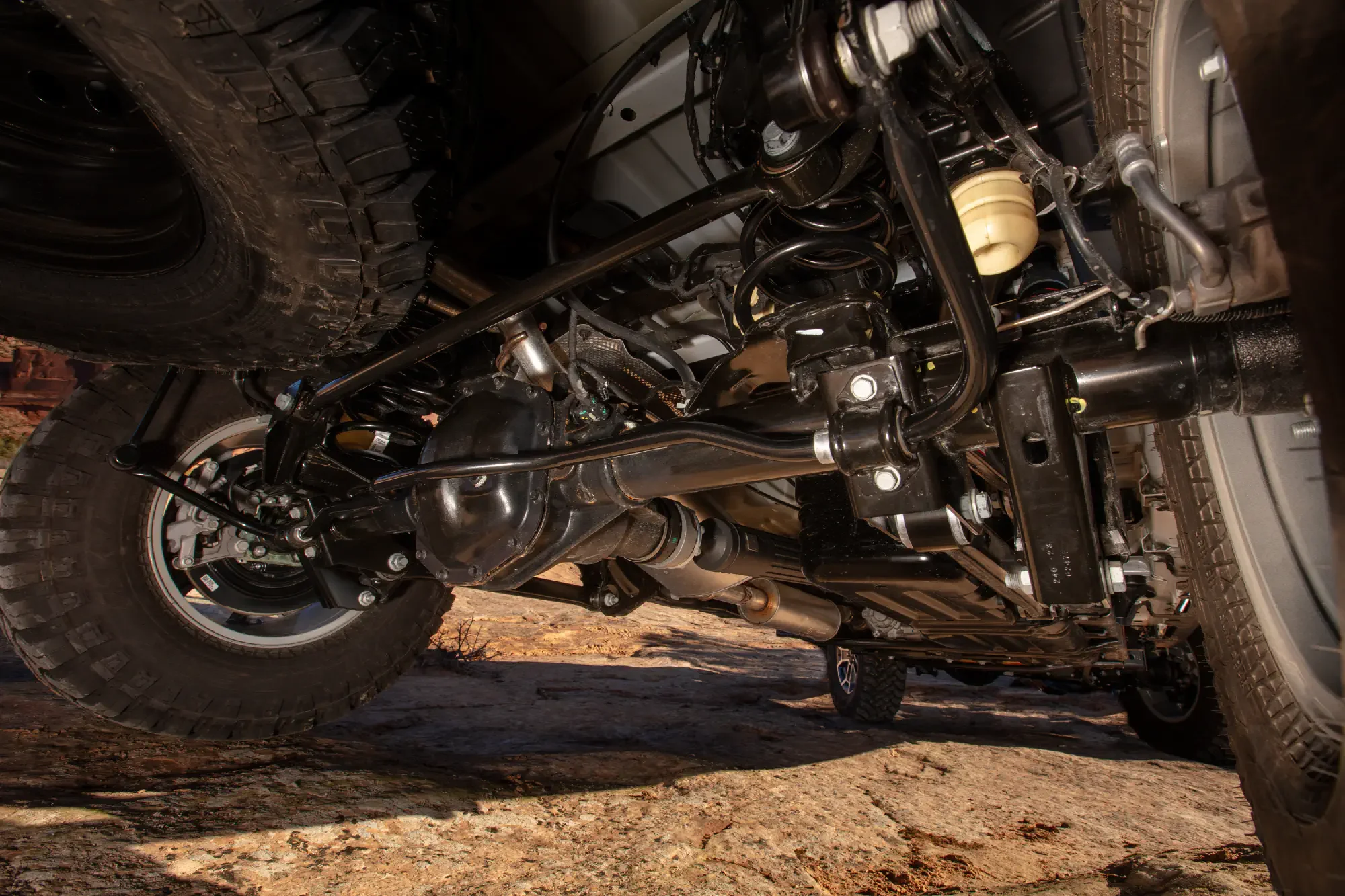 Underneath view of a vehicle showing the suspension system, tires, and parts of the drivetrain, with a dirt or gravel surface below and a brick wall in the background.
