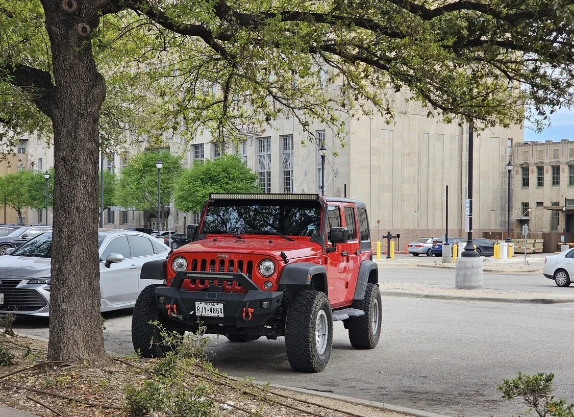 A red Jeep Wrangler parked on the side of a street near some trees and other parked cars, with buildings in the background.