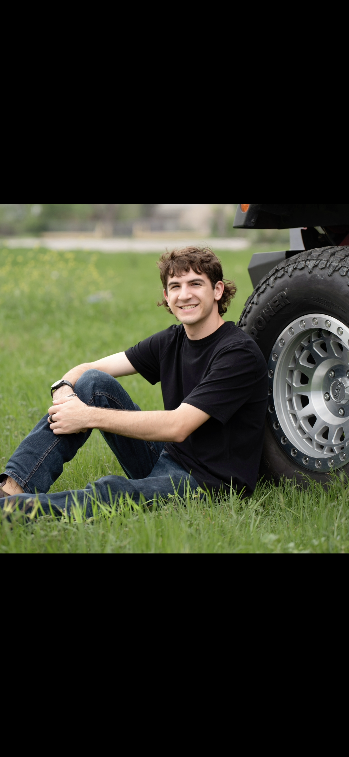 Young man sitting on grass next to a large vehicle tire, smiling at the camera in an open grassy field with trees in the background.