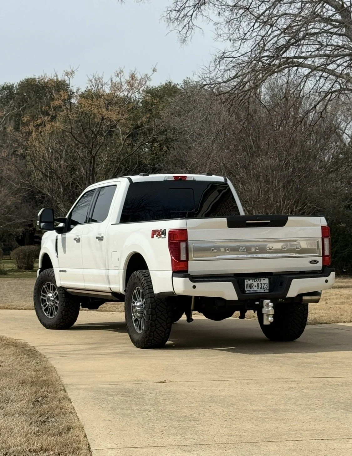 White lifted Ford F-150 truck with large off-road tires parked on a concrete driveway, with bare trees and grass in the background.