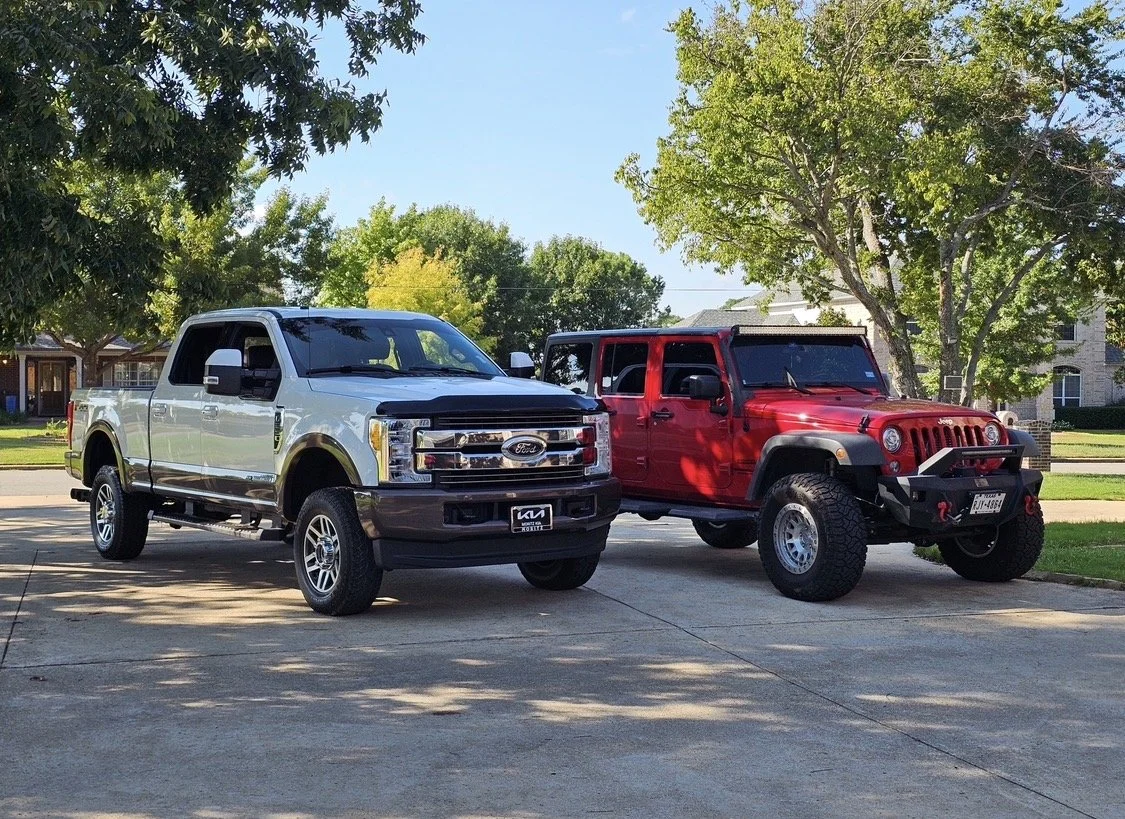 A white and black Ford truck and a red Jeep Wrangler parked on a driveway under trees in a suburban neighborhood.