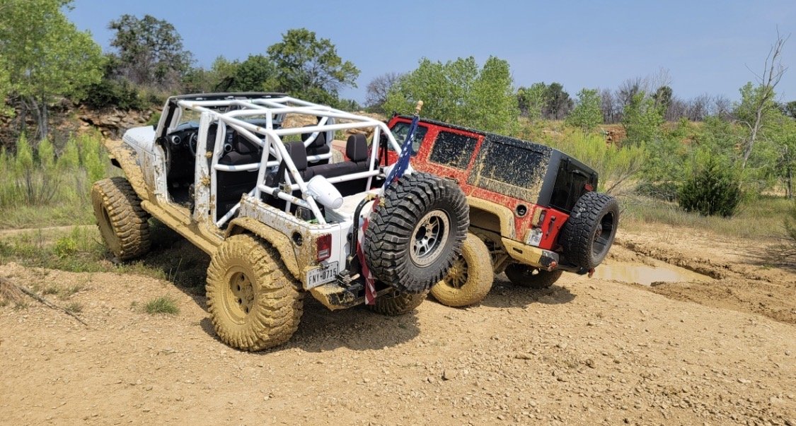 Two off-road vehicles, one white and one red, stuck in a muddy, uneven terrain during daytime with a clear blue sky and green trees in the background.