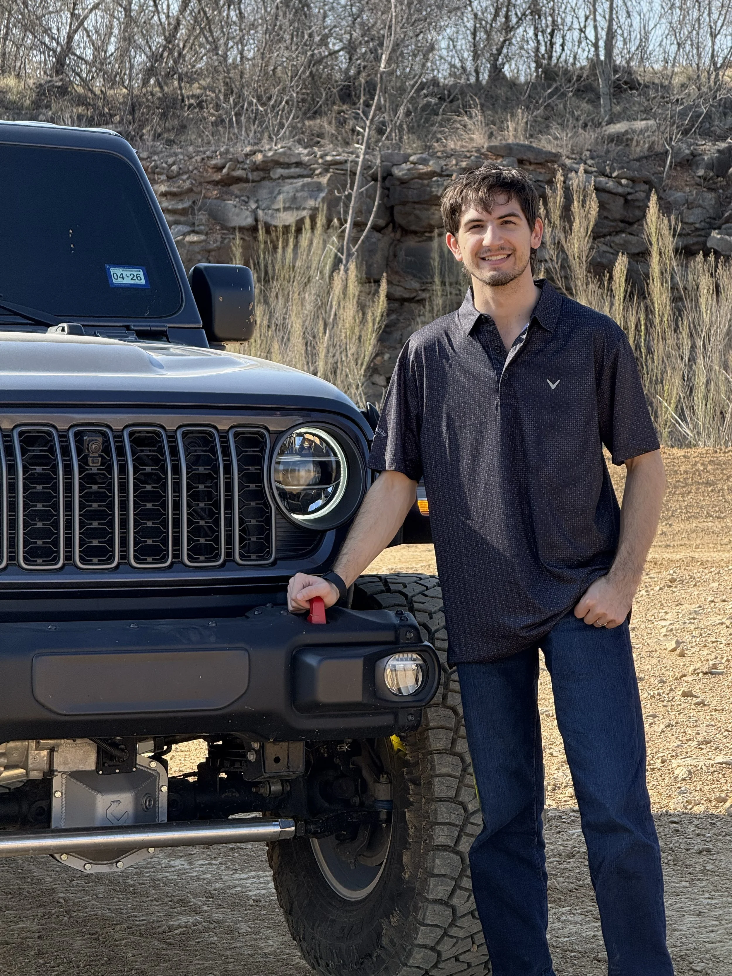 A young man with dark hair and a goatee wearing a black short-sleeved shirt and jeans standing outdoors on a dirt road beside a black Jeep vehicle in a desert landscape with rocks, dry bushes, and leafless trees.
