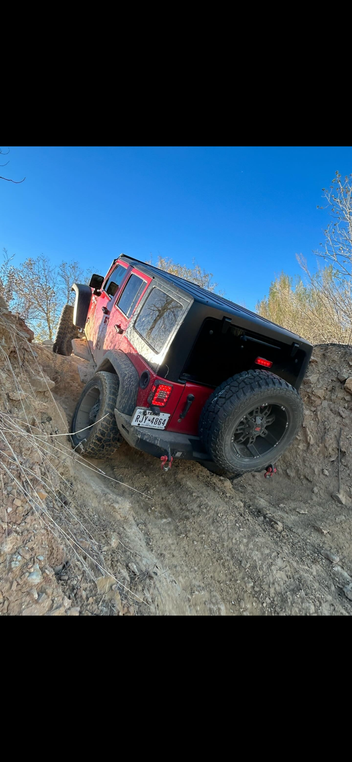 A red and black Jeep off-roading on a rugged dirt trail with steep incline and sparse trees in the background.