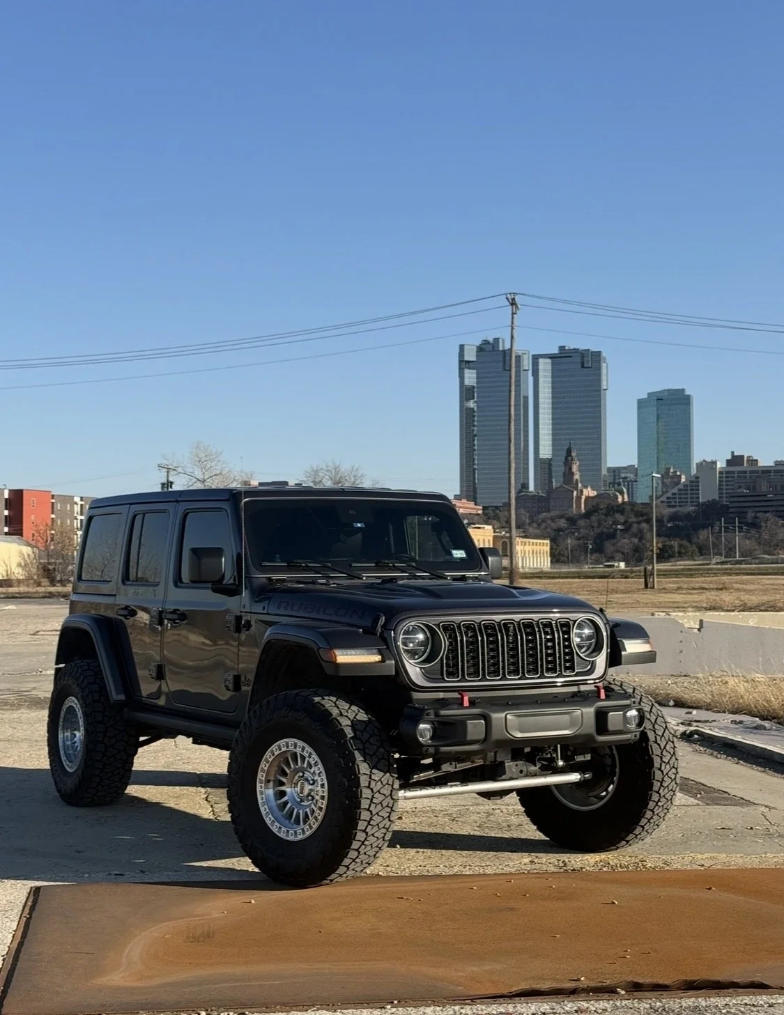 A black Jeep Wrangler parked outdoors with a city skyline in the background, under a clear blue sky.