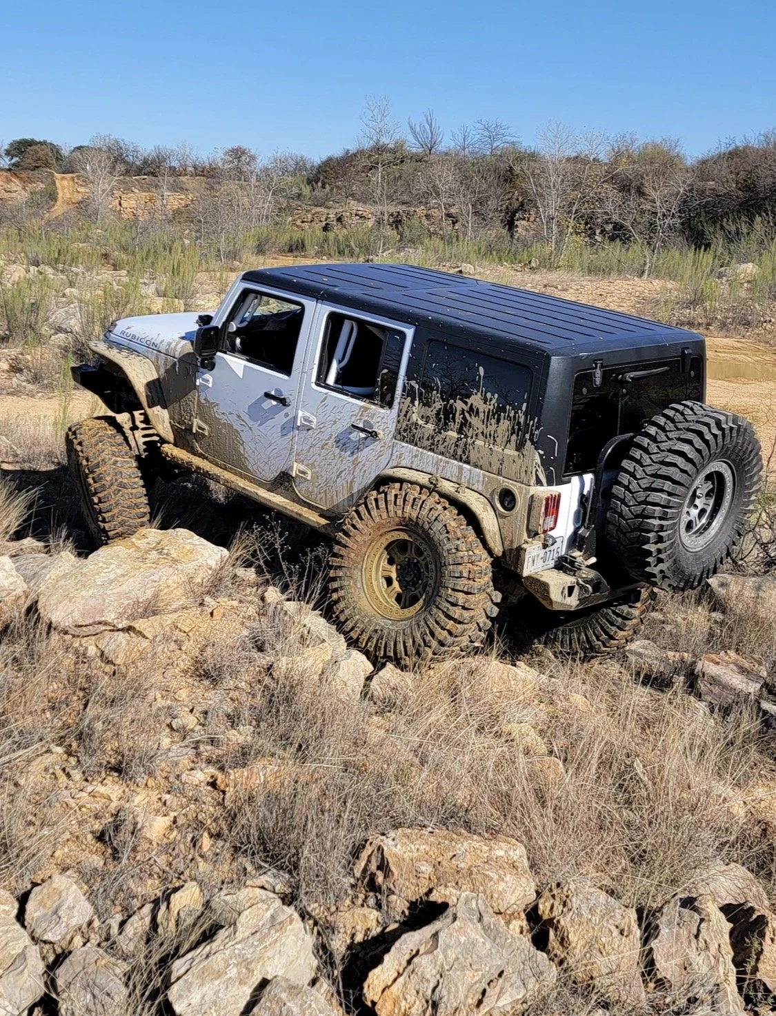 A four-door off-road vehicle climbing over rocks in a dry landscape with sparse vegetation and a clear blue sky.