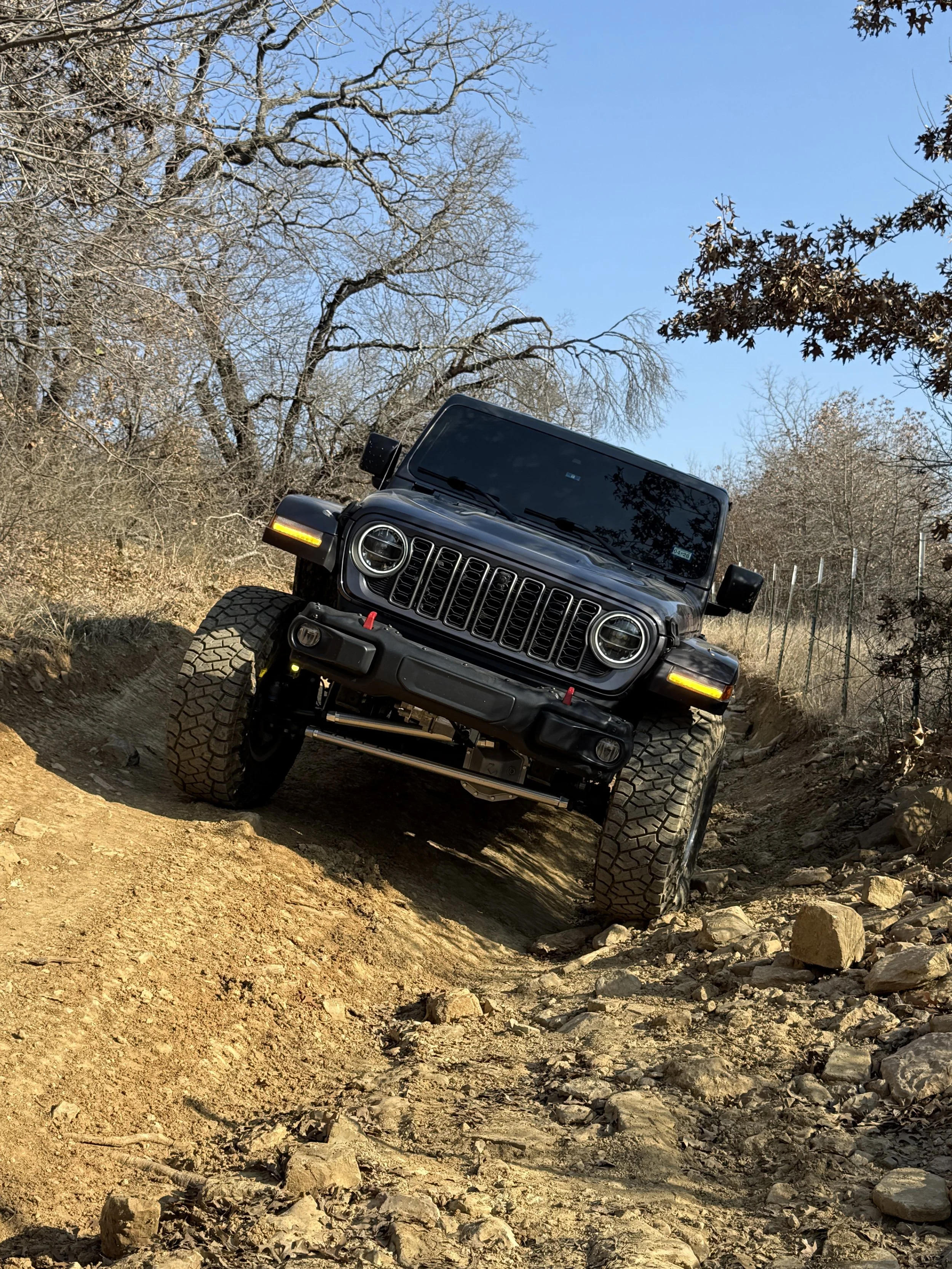 A black Jeep off-road vehicle driving on a rocky trail through a wooded area with leafless trees, in a dry landscape under a clear blue sky.