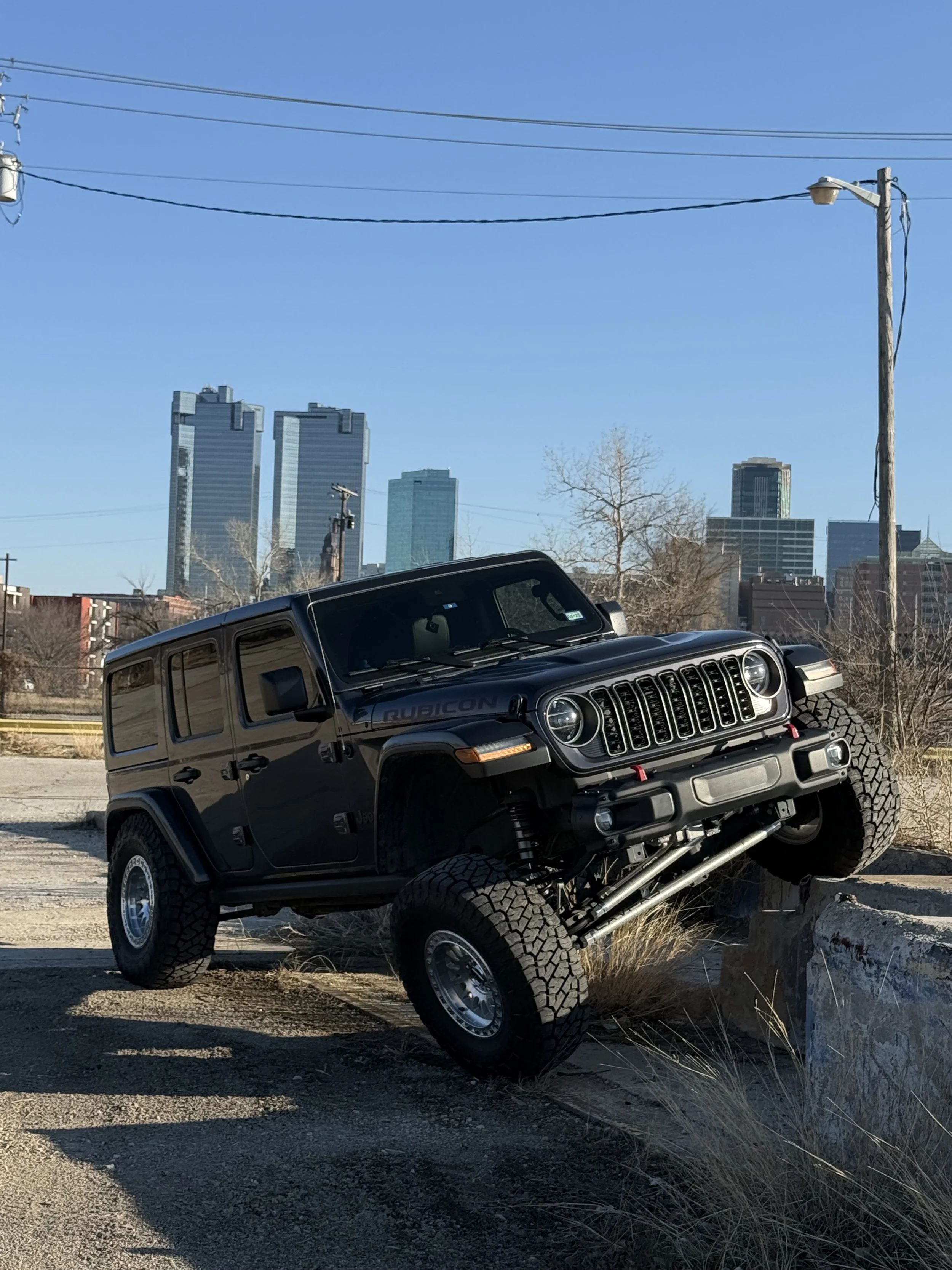 A black Jeep Rubicon with large off-road tires, driving over a concrete barrier with one front wheel elevated. The background shows a city skyline with tall skyscrapers and some leafless trees, under a clear blue sky.