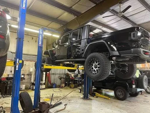 A black modified off-road vehicle is elevated on a hydraulic lift inside an automotive repair shop with various tools and equipment visible around.