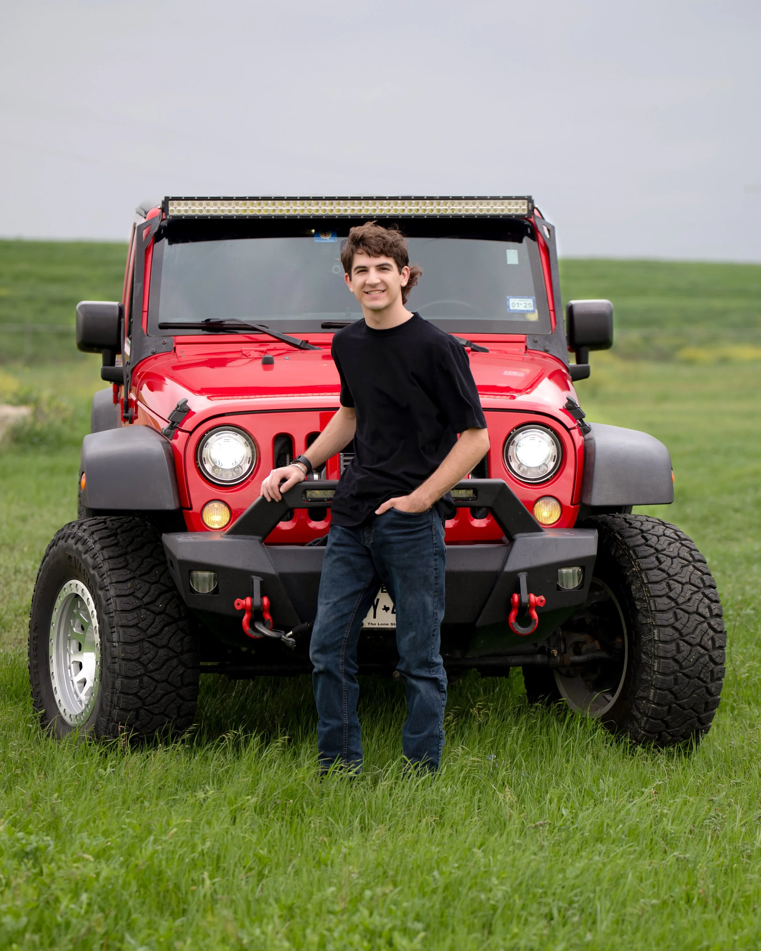 A young man in a black t-shirt and jeans standing in front of a red Jeep Wrangler on a grassy field, smiling at the camera.