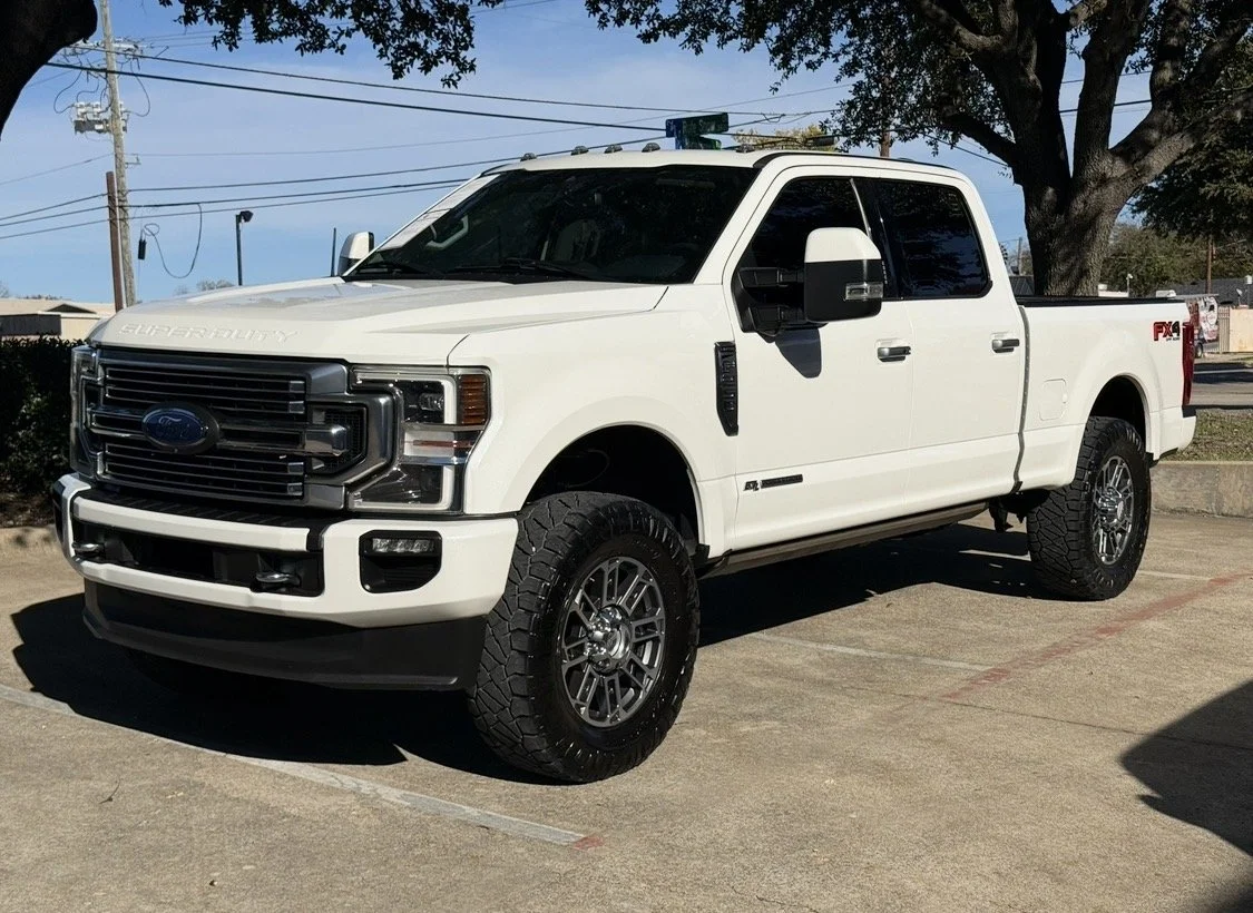 A white Ford Super Duty pickup truck parked on a concrete lot with trees and power lines in the background.