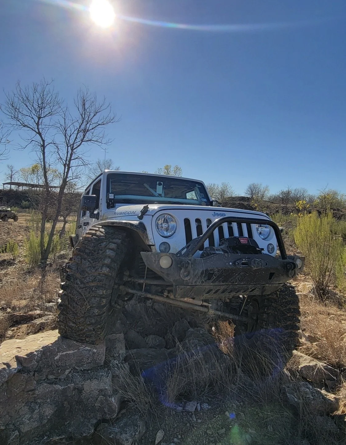 A white Jeep Rubicon off-road vehicle climbing over rocks in a dry, desert-like landscape on a sunny day.