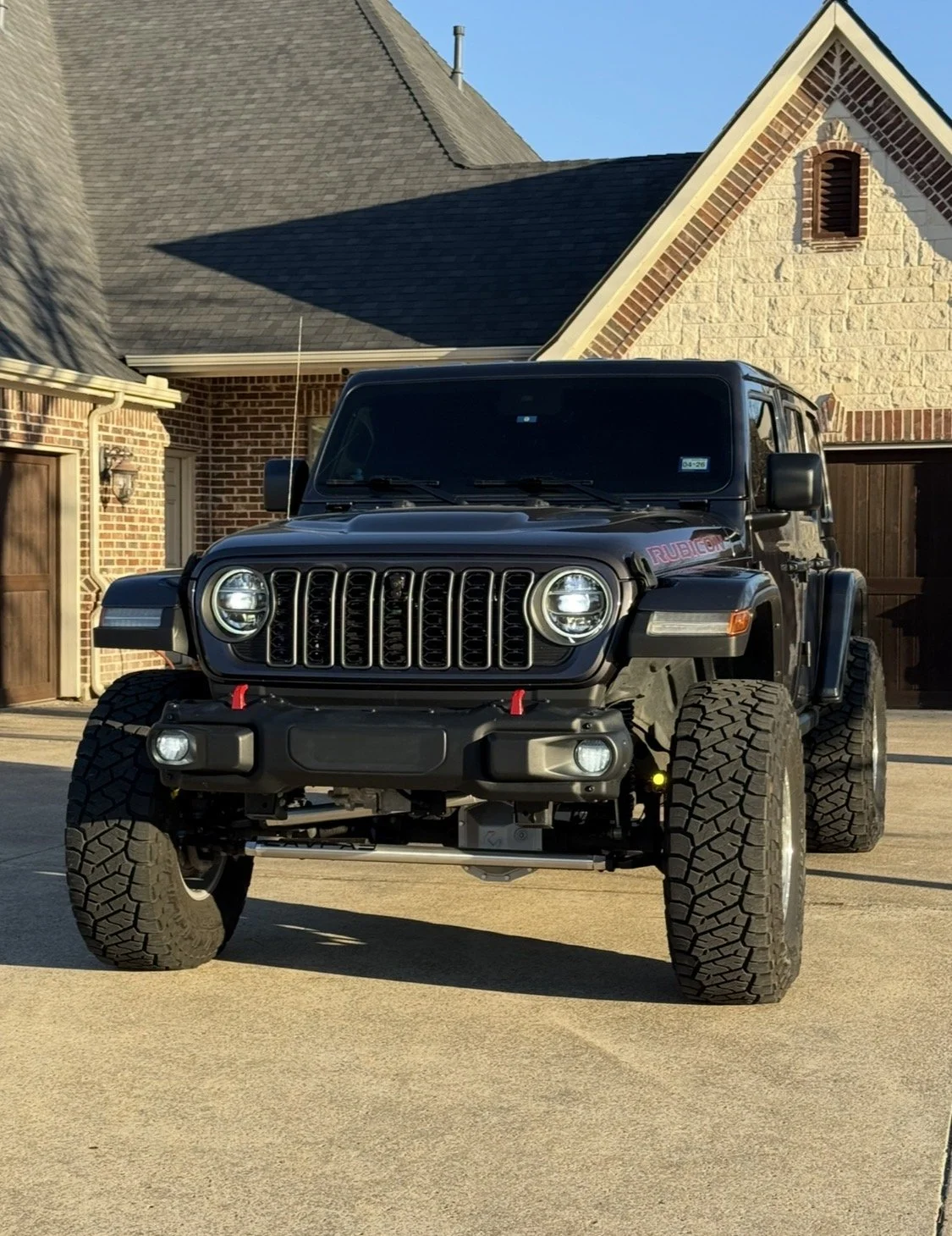 Front view of a black Jeep Rubicon SUV parked on a driveway in front of a brick house with a pitched roof.