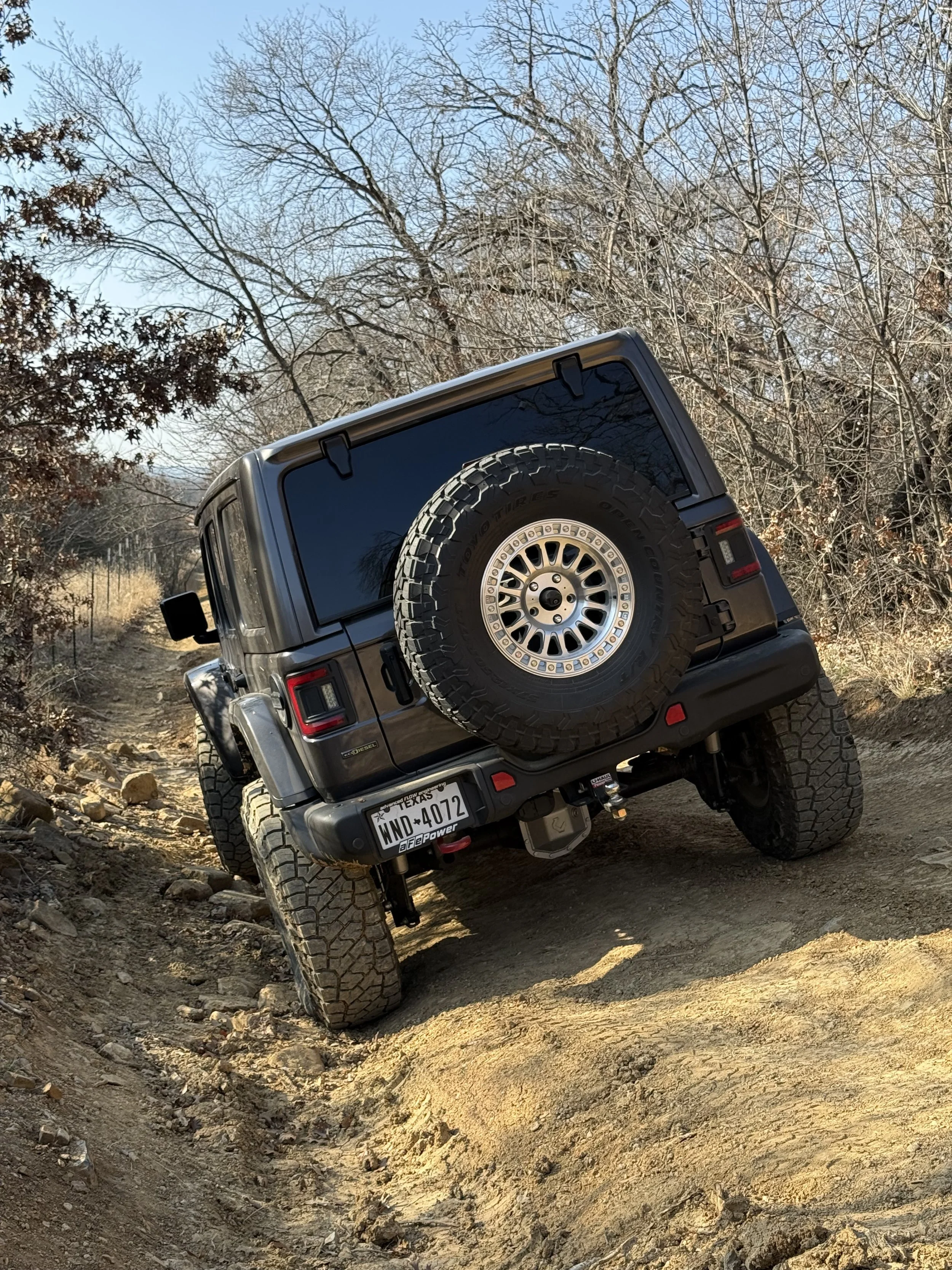 A black Jeep Wrangler off-road vehicle navigating uneven terrain on a dirt trail surrounded by leafless trees.