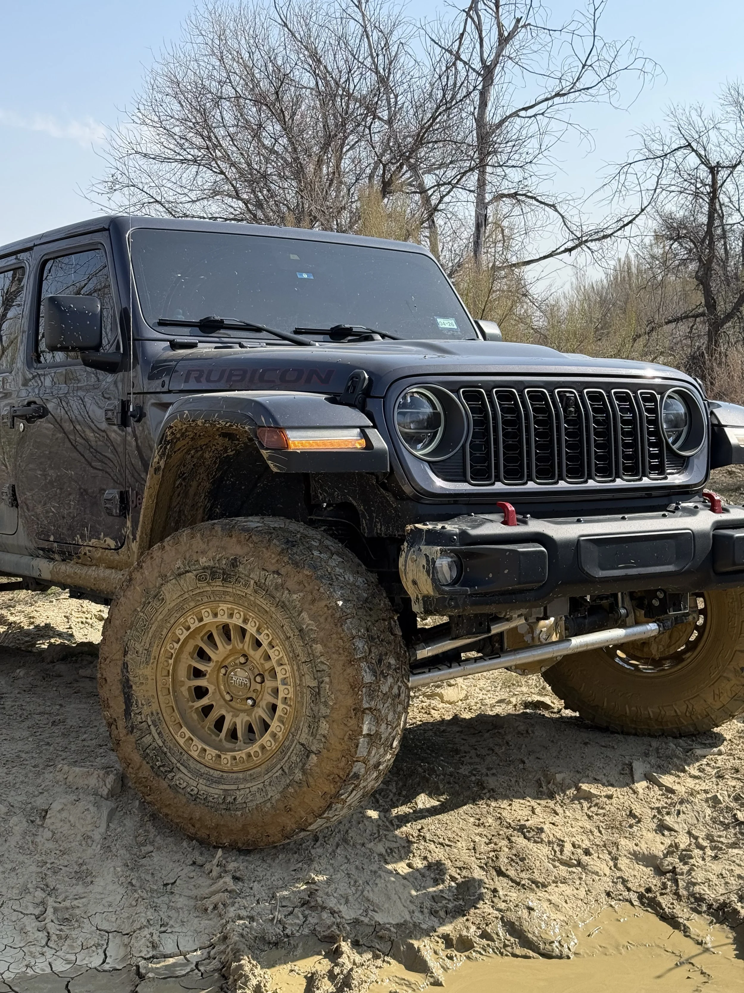Black Jeep Rubicon off-road vehicle with muddy tires on dirt ground, surrounded by leafless trees under a partly cloudy sky.