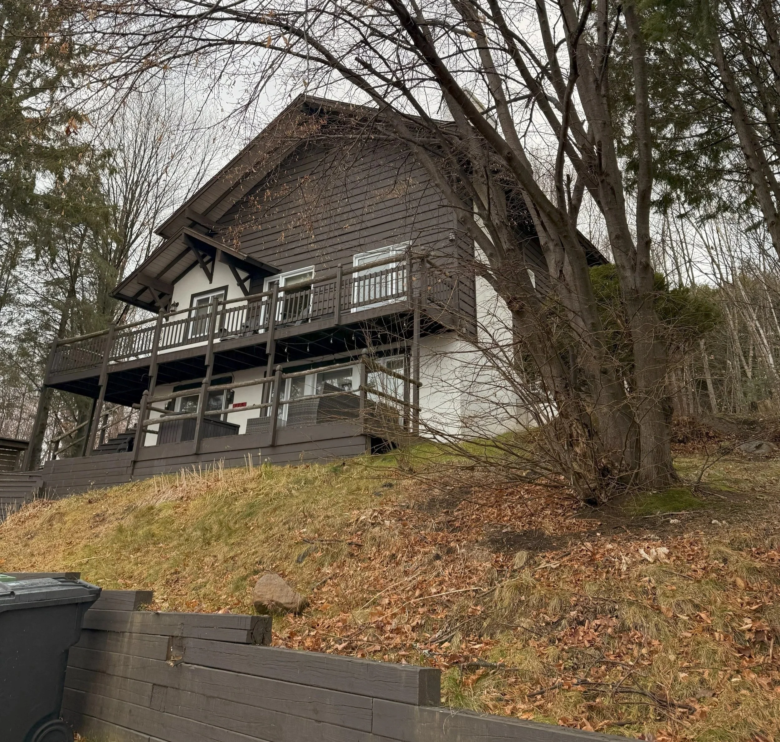 Maison en bois avec plusieurs étages, située sur une pente, entourée d'arbres nus, avec une terrasse en bois à l'avant. Le ciel est overcast.